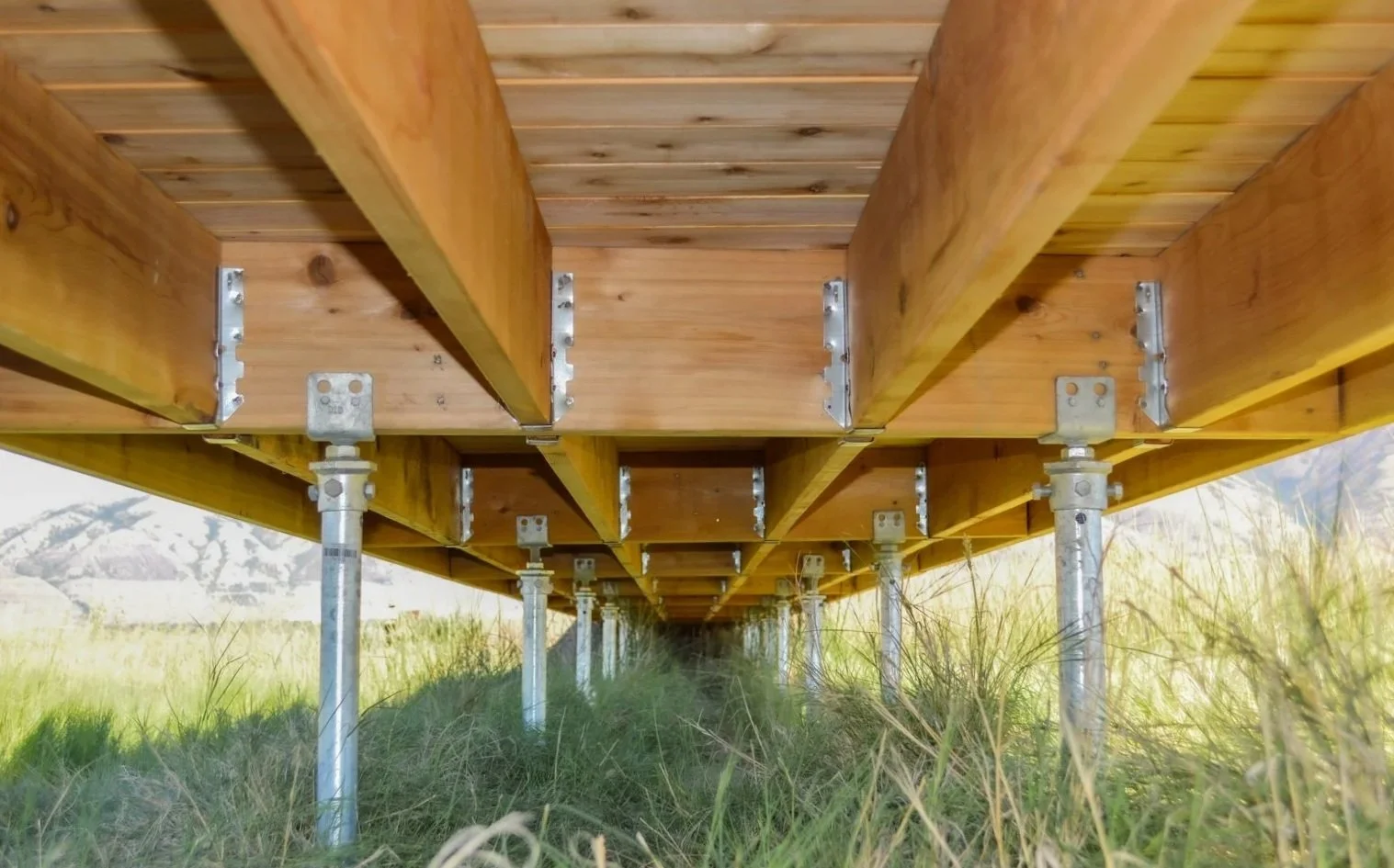 Underside of a wooden deck with support beams and metal poles, set in grassy field with mountains in the background.