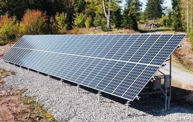 Solar panel array installed outdoors on a gravel surface with trees and foliage in the background. Ground-mounted solar panel foundation installed with helical piles in Western New York.