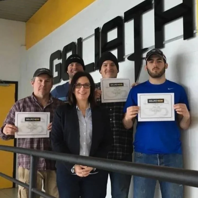 A group of five people standing indoors in front of a white wall with large black letters, holding certificates, and smiling.