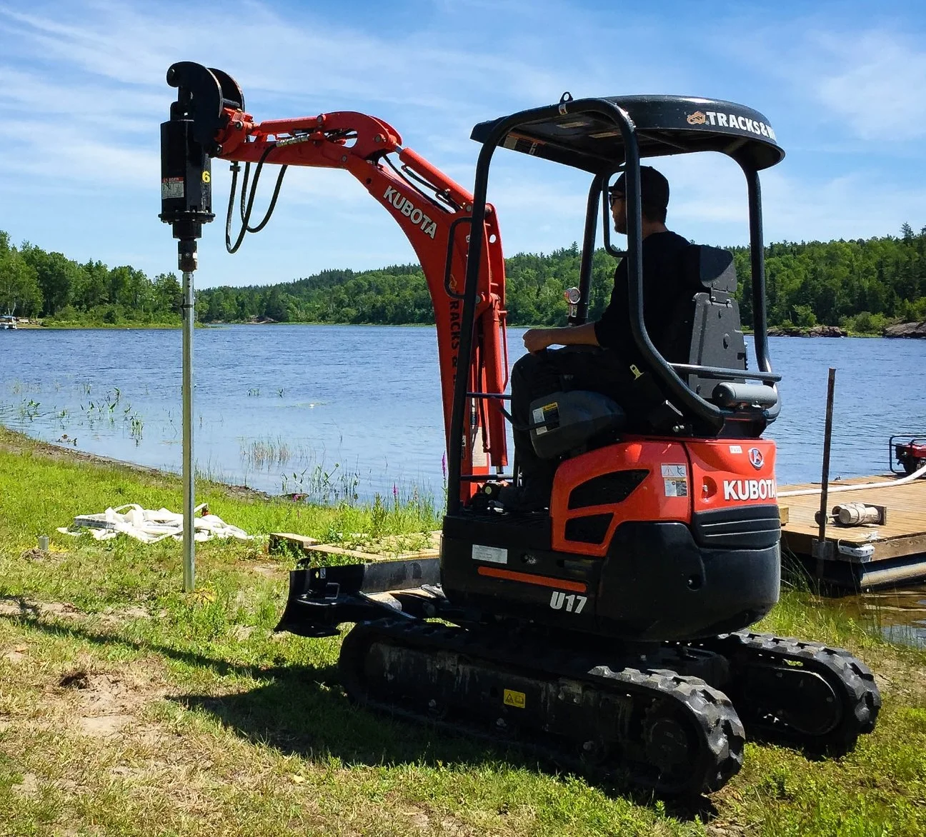 A mini excavator with a red arm and black body parked on grass near a lake with a dock and a wooded shoreline, under a partly cloudy sky.