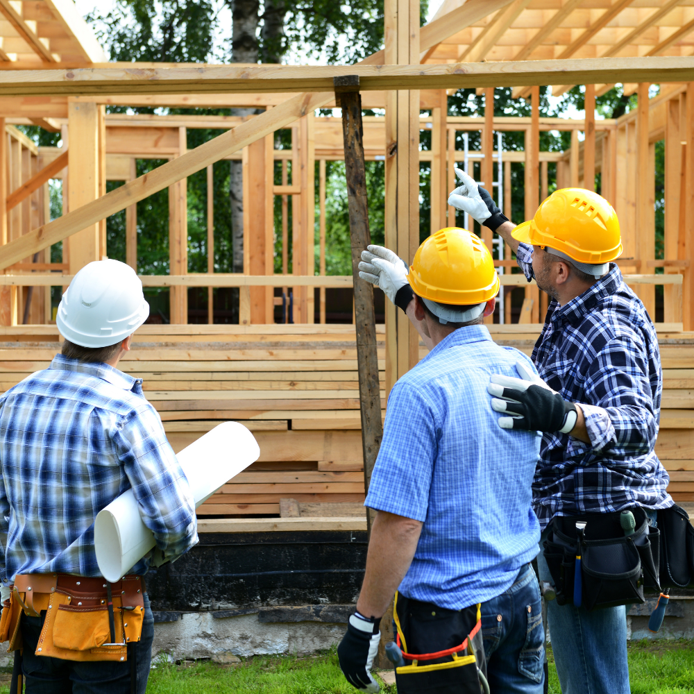 Three construction workers wearing safety helmets and gloves at a building site, discussing the framing of a wooden house under construction.