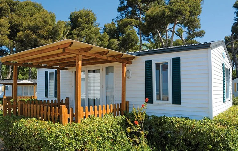 White mobile home with black shutters, a small wooden porch and rain protection roof, surrounded by green bushes and trees under a clear blue sky.