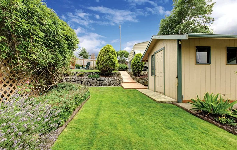 A well-maintained backyard with lush green grass, flower beds, a garden shed, and a paved walkway, with trees and shrubs in the background under a partly cloudy sky.