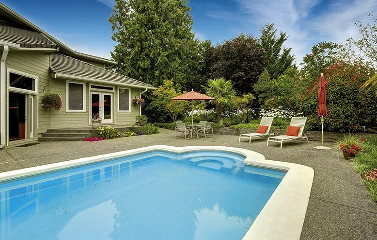 A backyard with a swimming pool, lounging chairs with orange cushions, a table with an umbrella, and surrounding greenery and plants.