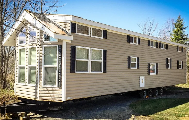 A beige mobile home with multiple windows and black shutters, situated on a trailer foundation on a gravel lot, with trees and a clear blue sky in the background.