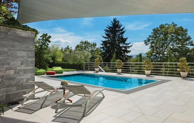 Outdoor patio with a swimming pool, two lounge chairs, potted plants, green trees, and a clear blue sky.