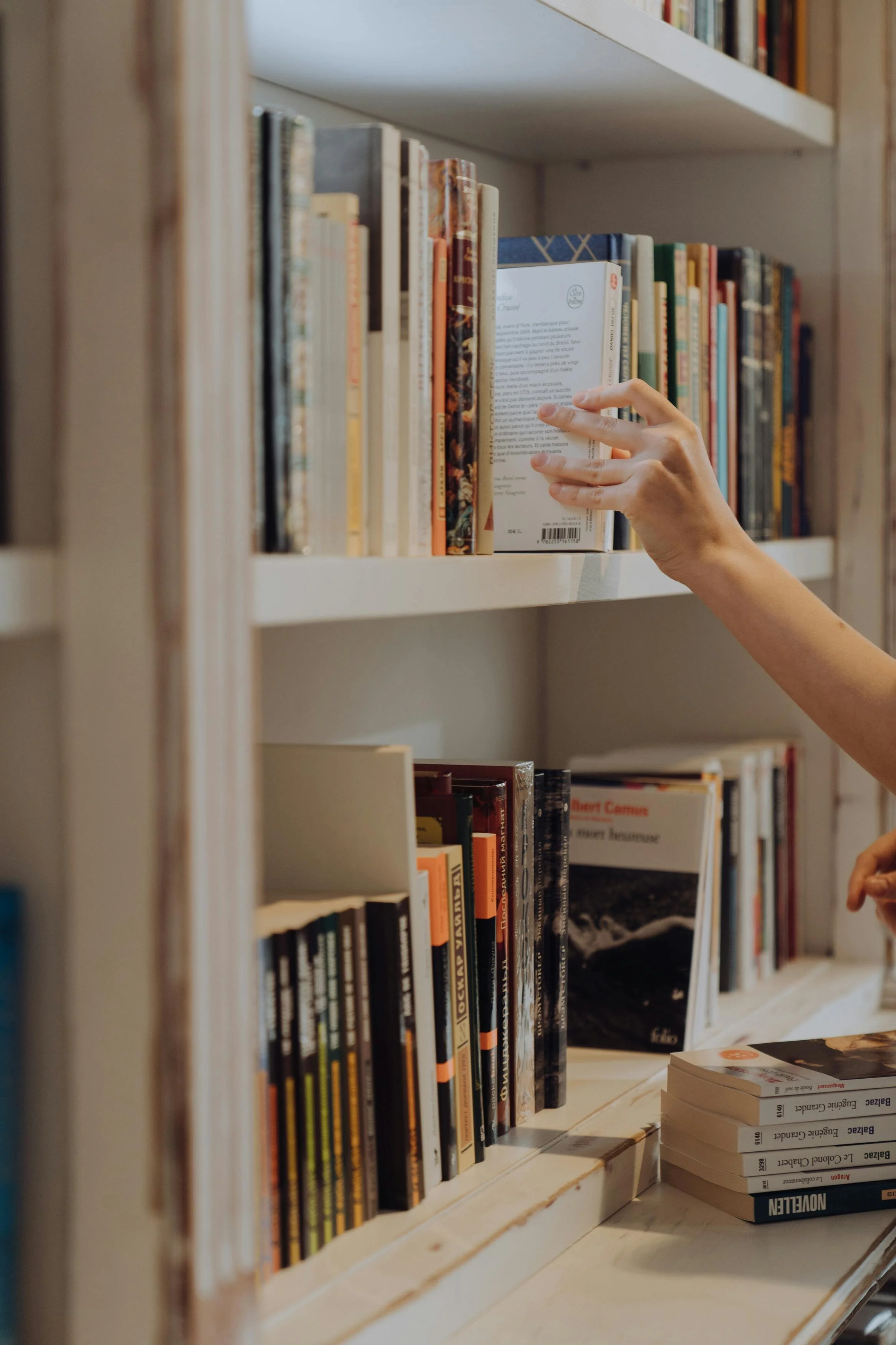 A person reaching out to grab a book on a white bookshelf filled with books in a bookstore or library.