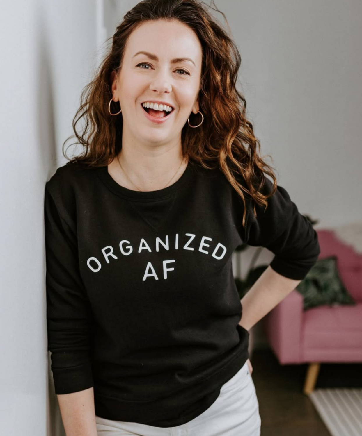 Kelle wearing a black sweatshirt with white text that reads 'ORGANIZED AF.' She is smiling, and leaning against a wall in a room with a pink armchair and green pillows in the background.