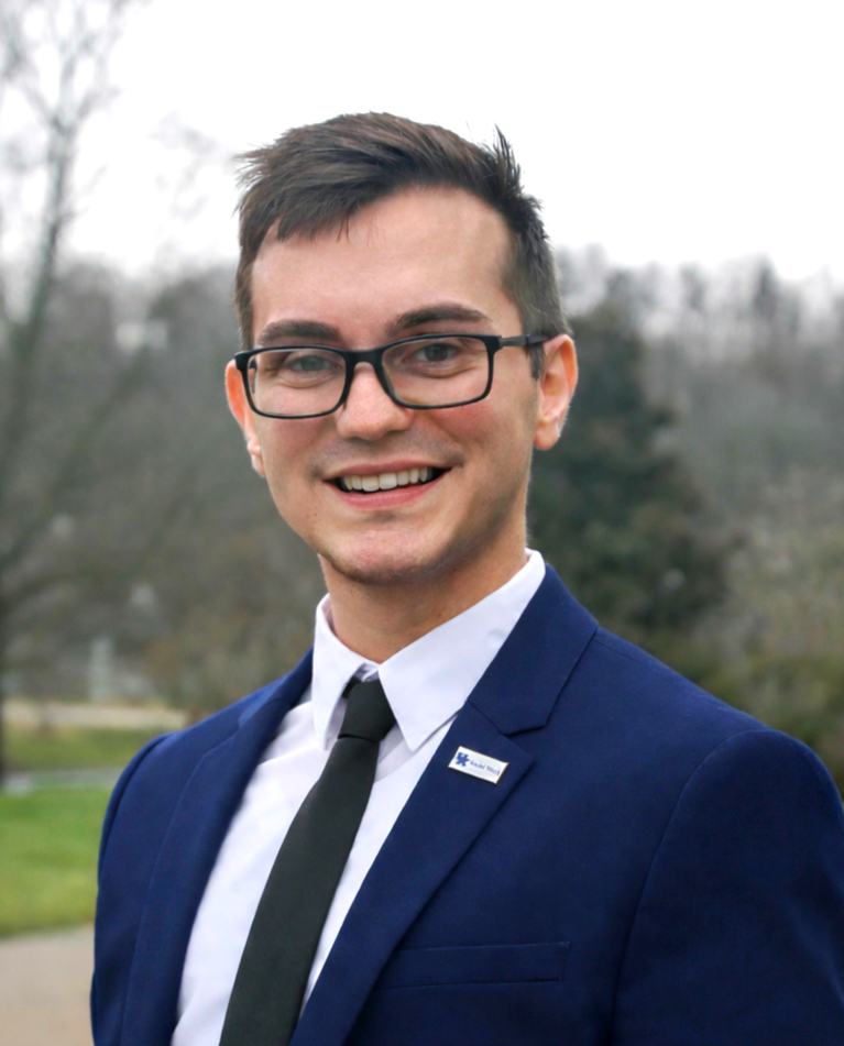 A young man in glasses, wearing a navy suit with a white shirt and black tie, smiling outdoors with trees in the background.