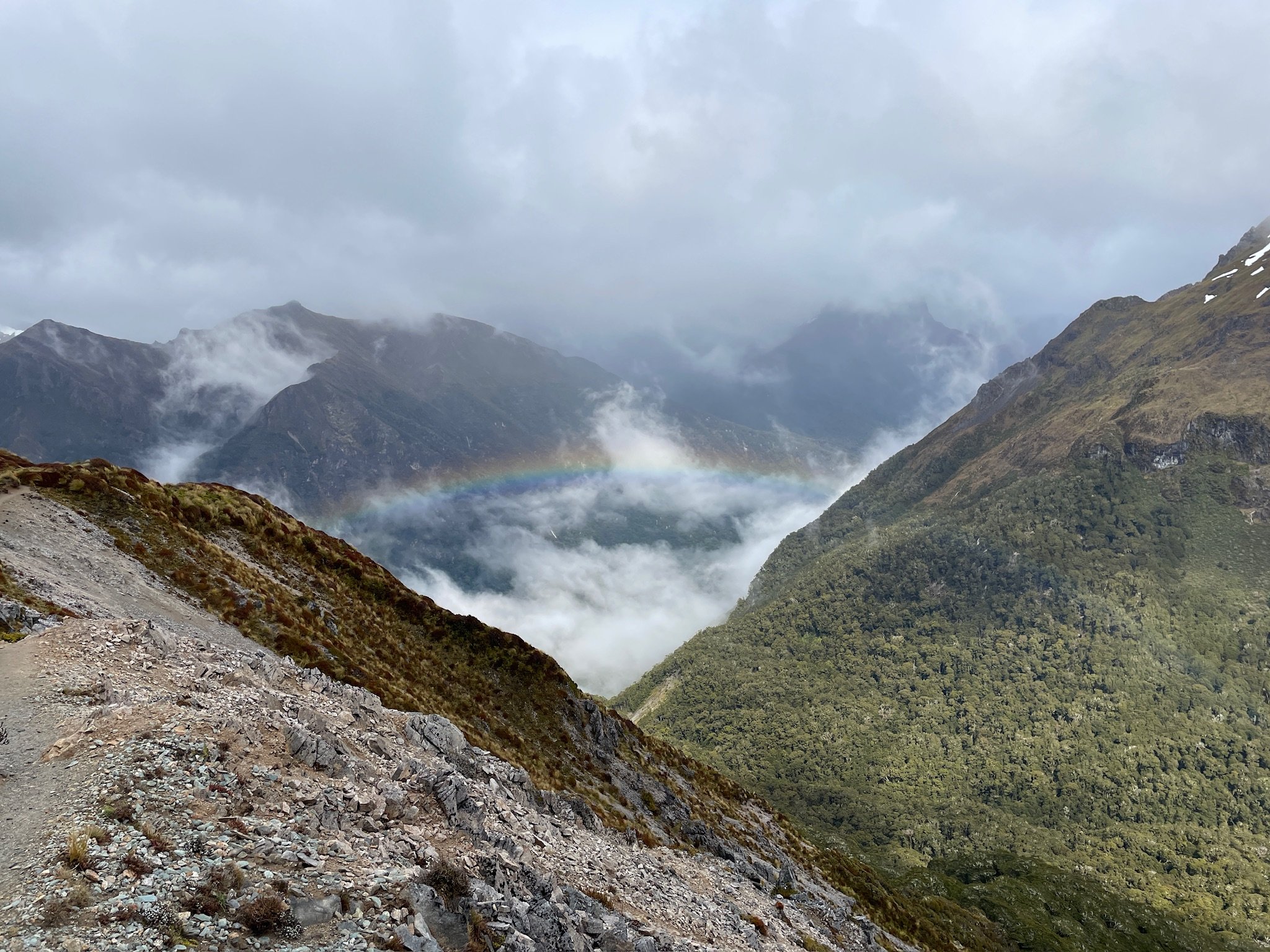 Mountain landscape with cloudy sky, a faint rainbow, and a rocky trail in the foreground.