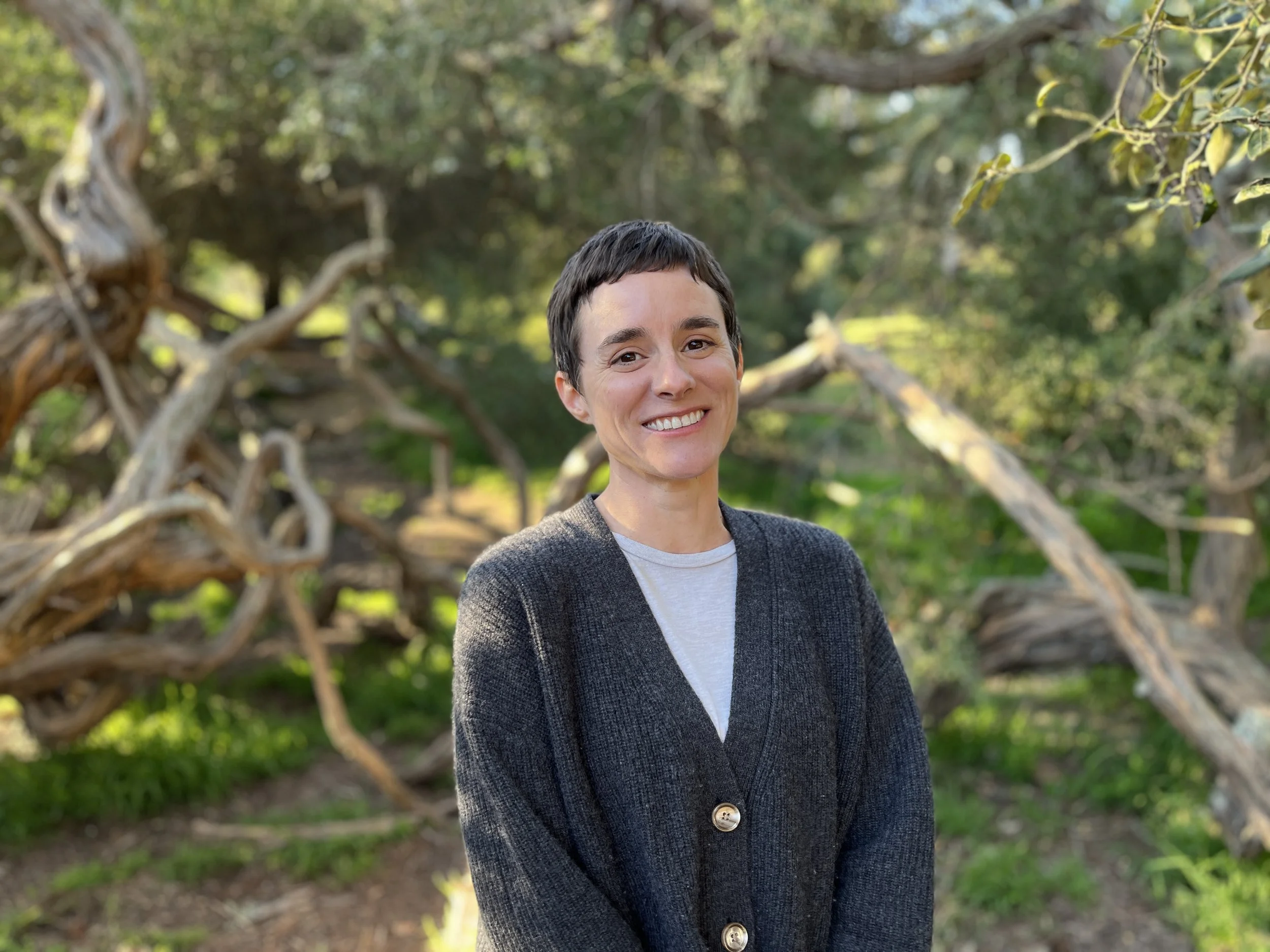 A woman with short dark hair smiling outdoors with green trees and twisted branches in the background.