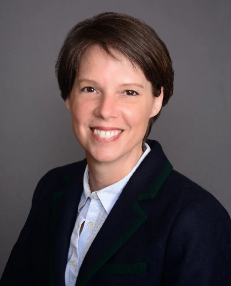 Professional woman with short brown hair, wearing a navy blazer and white shirt, smiling against a gray background.