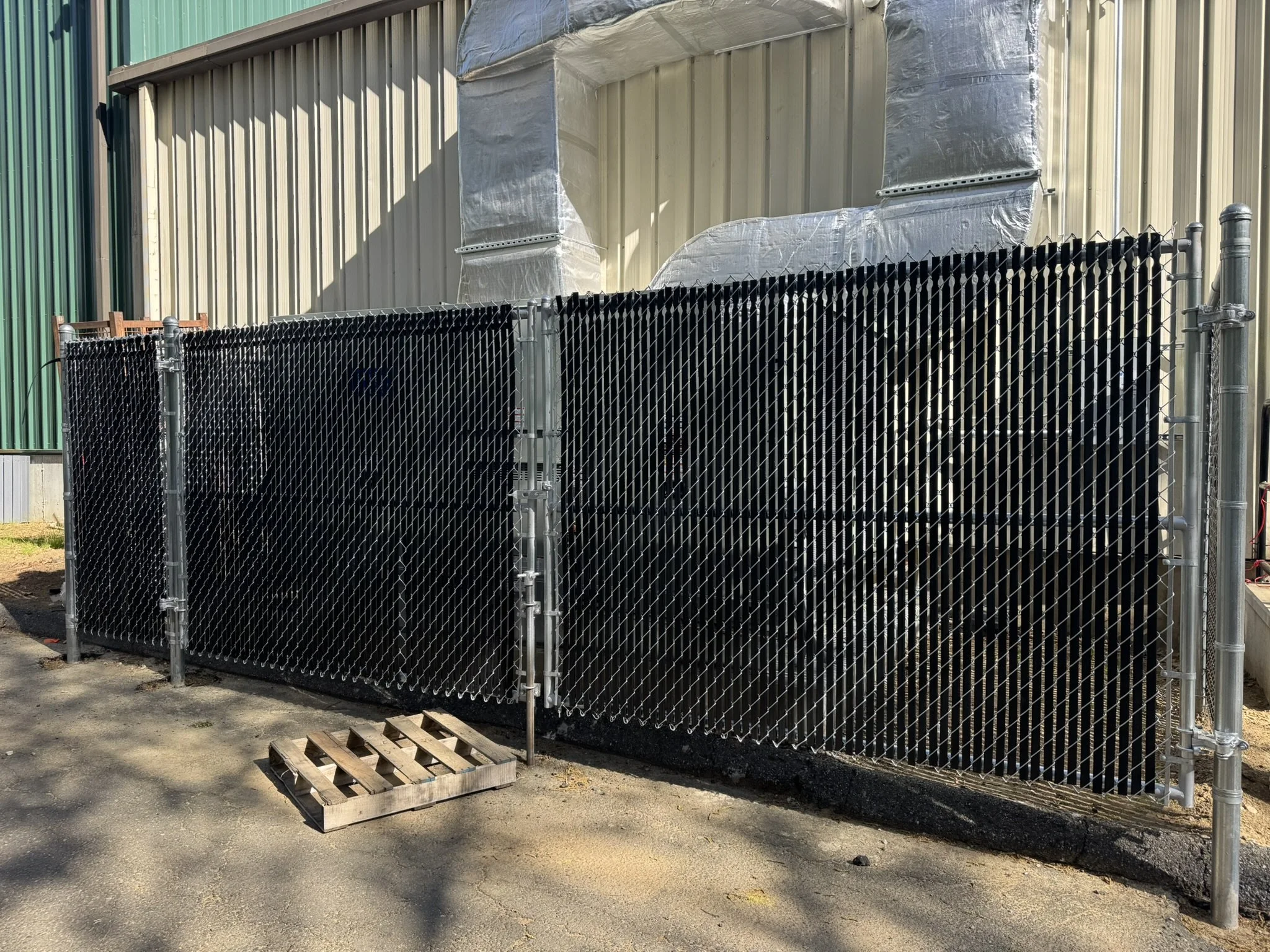 Black chain-link construction fence in front of a beige industrial building, with some shadow and a wooden pallet on the ground.