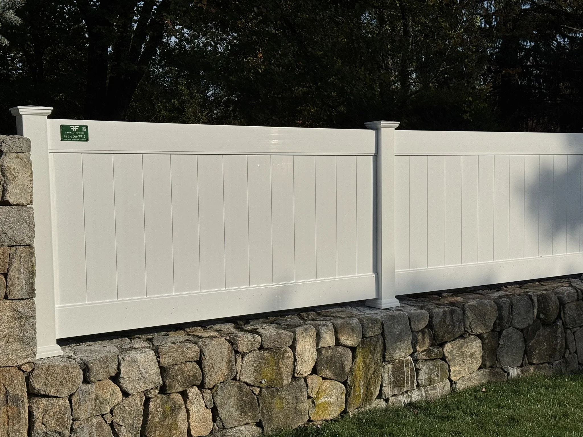 A white vinyl fence with stone base, running along a grassy area with trees in the background.