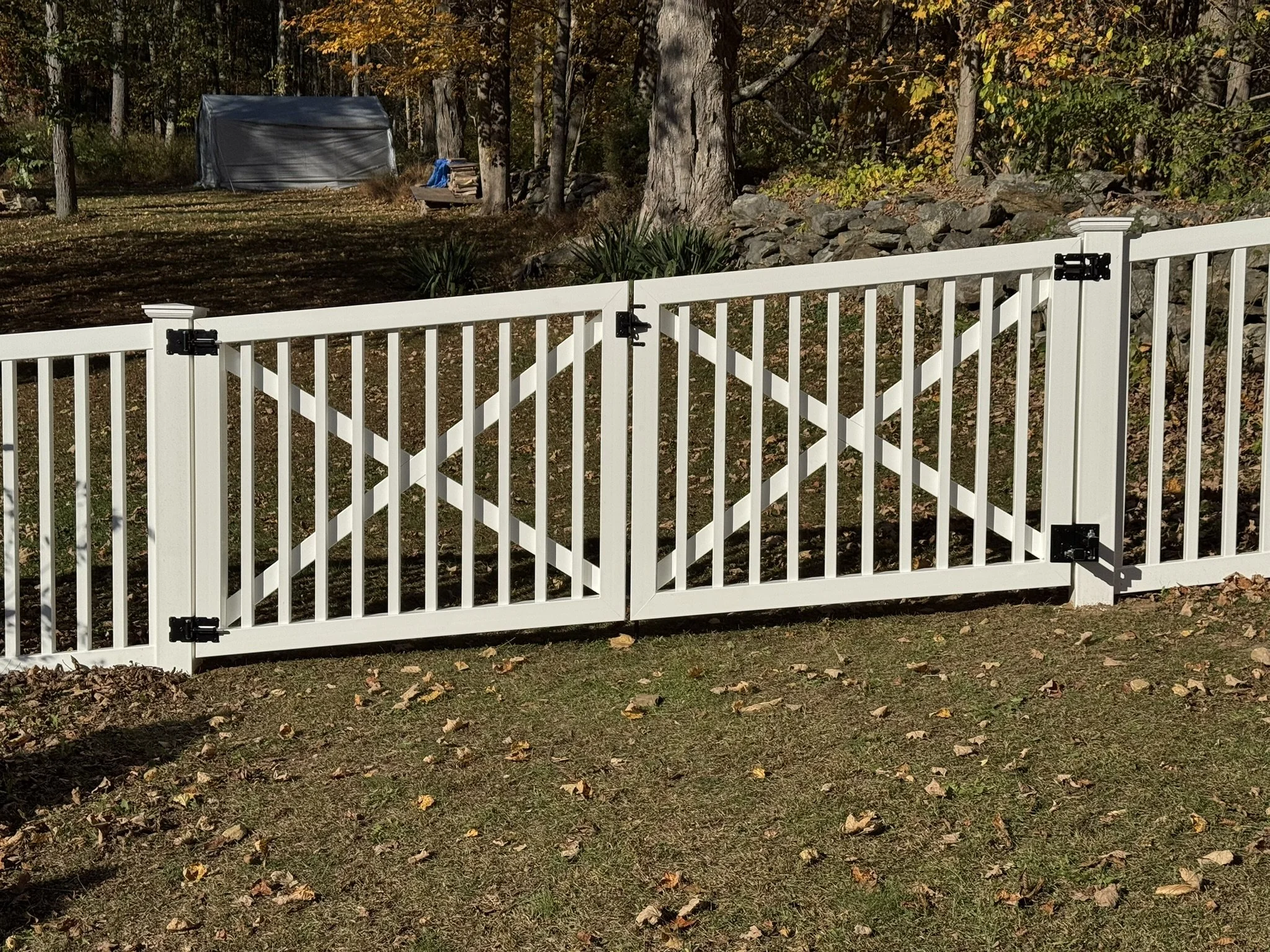 A white picket fence gate with black hinges, set in a yard with fallen leaves, trees, rocks, and a blue tarp or tent in the background.