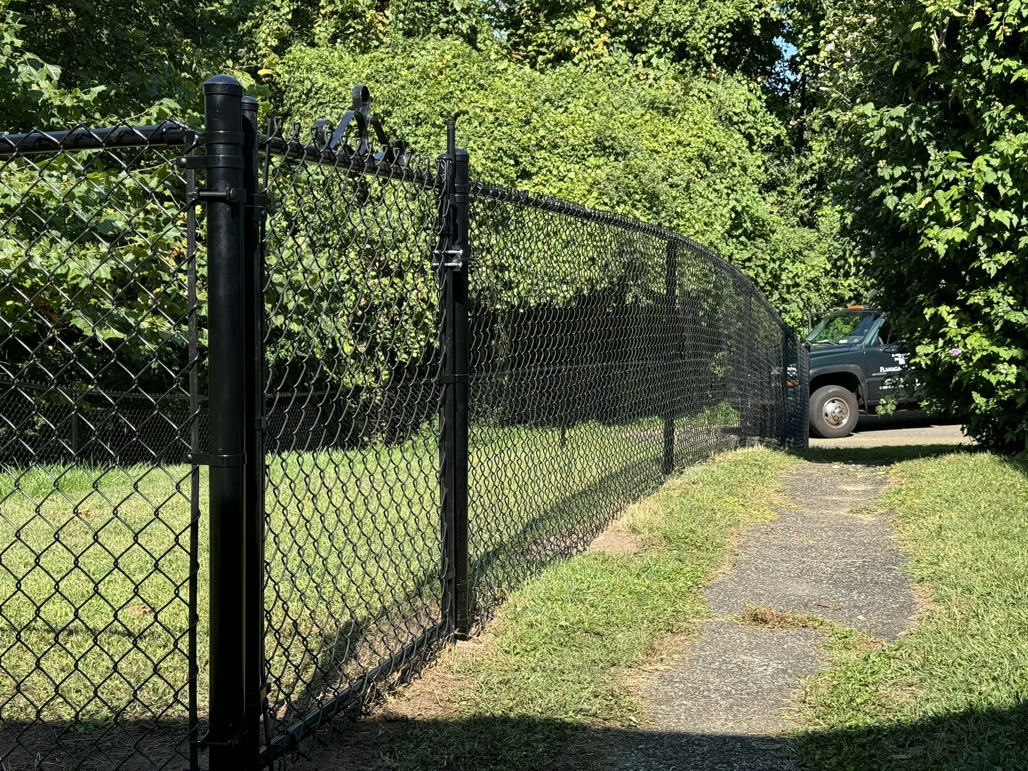 Black metal fence running along a sidewalk with greenery and trees in the background, and a dark-colored vehicle parked beyond the fence.