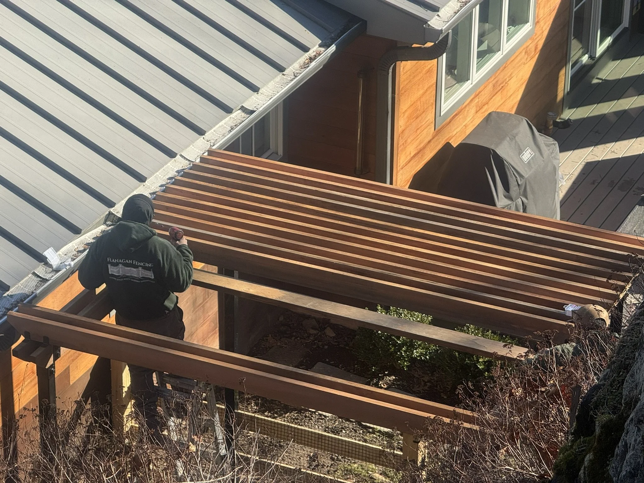 Worker installing wooden slats on a patio roof of a modern house with a wooden exterior and large windows.