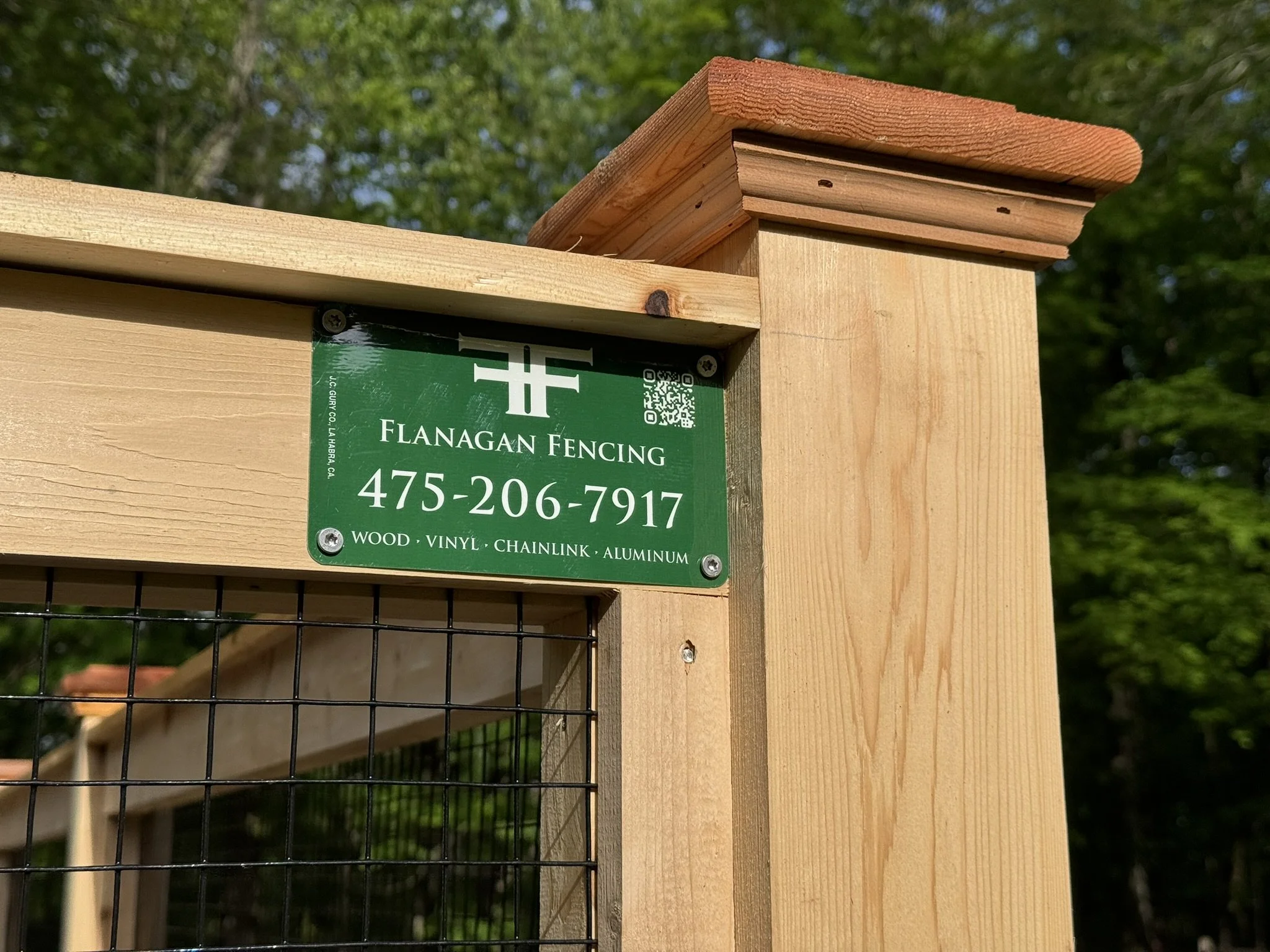 Close-up of a green sign with white text on a wooden fence post. The sign reads "Flanagan Fencing," includes a phone number, and lists materials such as wood, vinyl, chainlink, and aluminum. The fence is partially visible with a wire mesh.