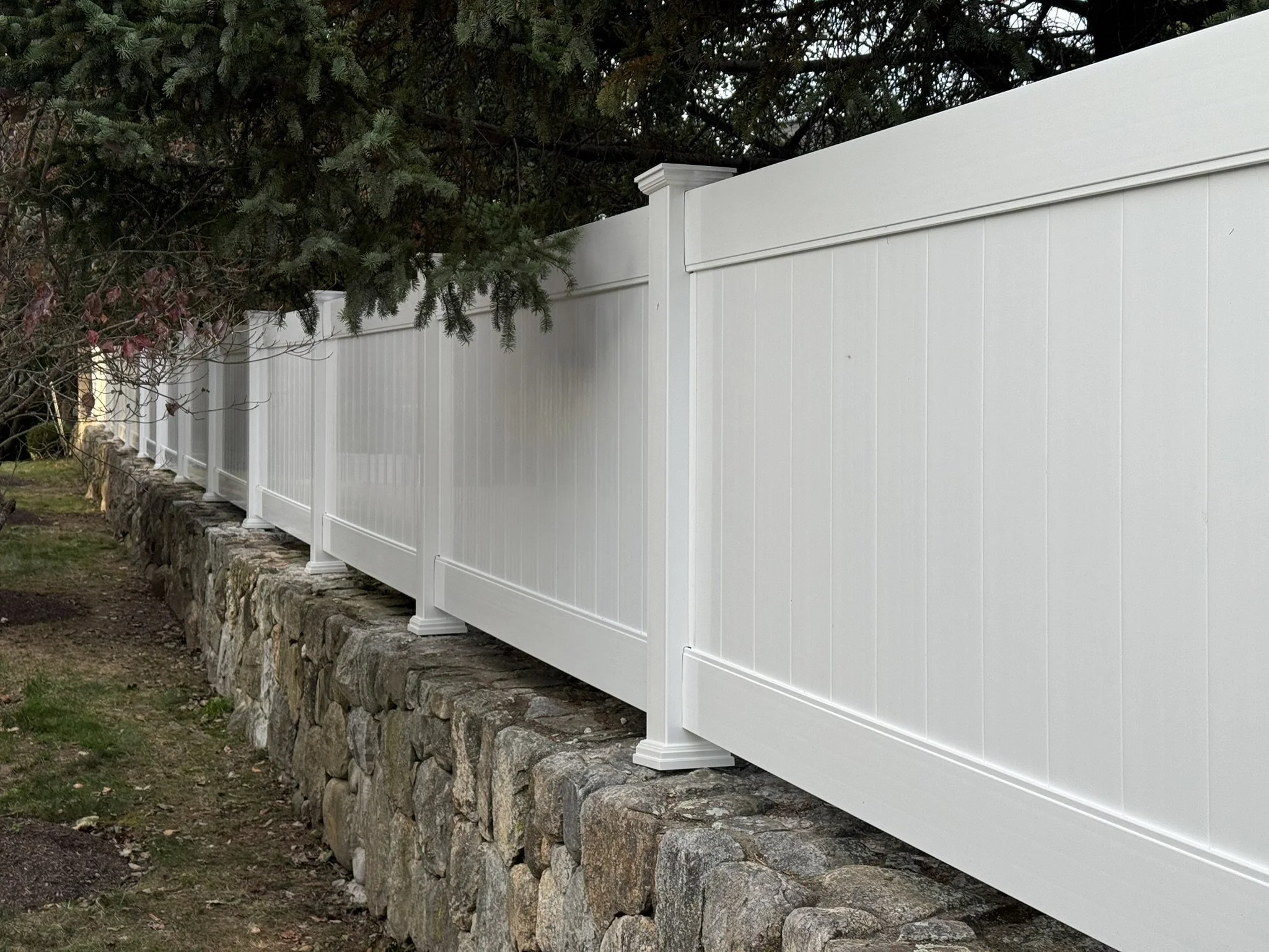 White vinyl privacy fence on a stone foundation, next to a grassy area and trees.