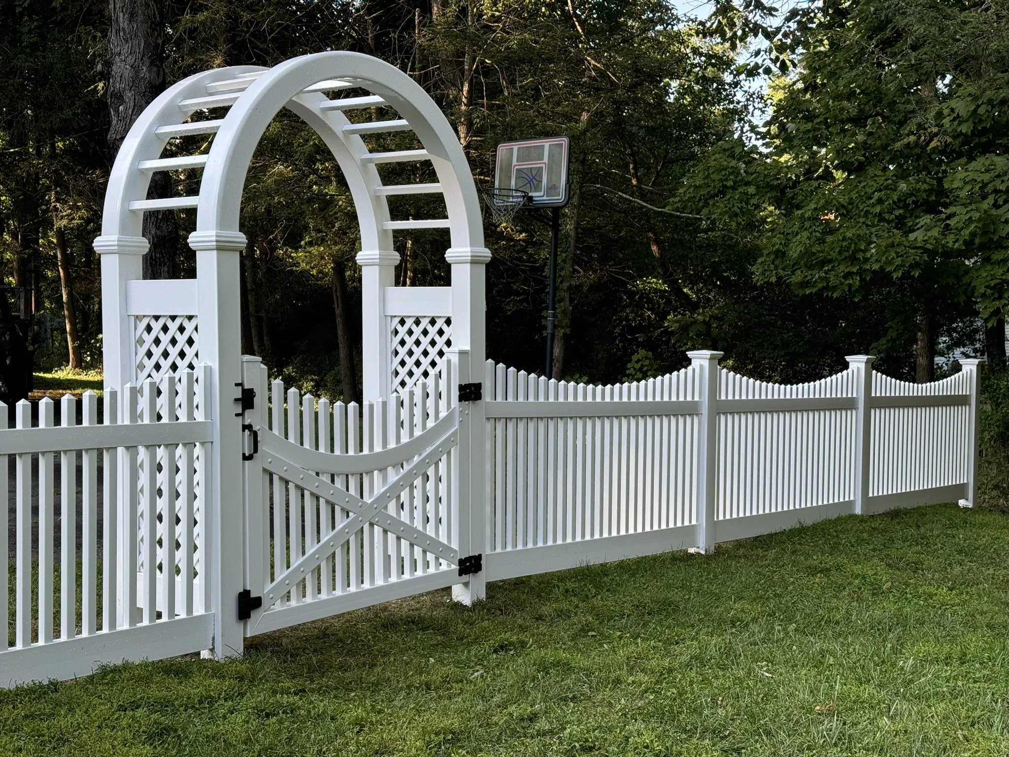 White picket fence with an arched gate, set on a grassy yard, with trees and a basketball hoop in the background.
