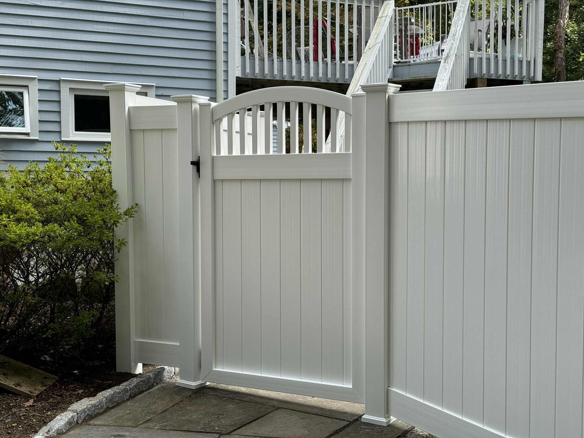 White vinyl privacy fence with a gate, adjacent to a landscaped garden with shrubs, in front of a blue house with small windows and a porch with stairs.