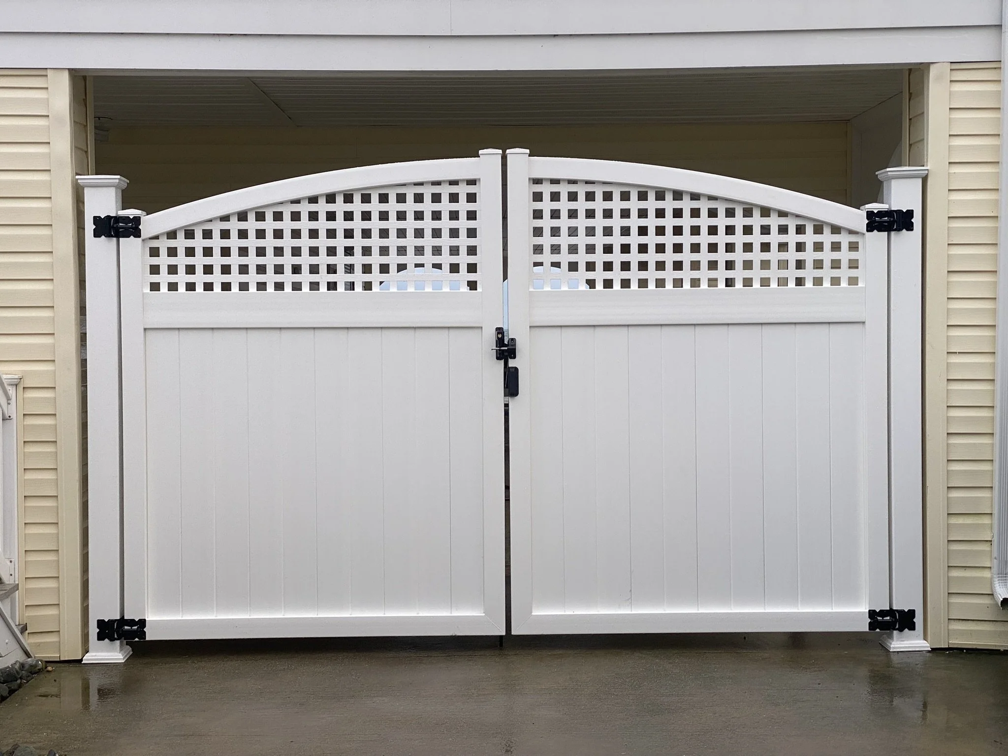 White fence gate with lattice top, black hardware, closed, in front of Beige house with siding, concrete ground.