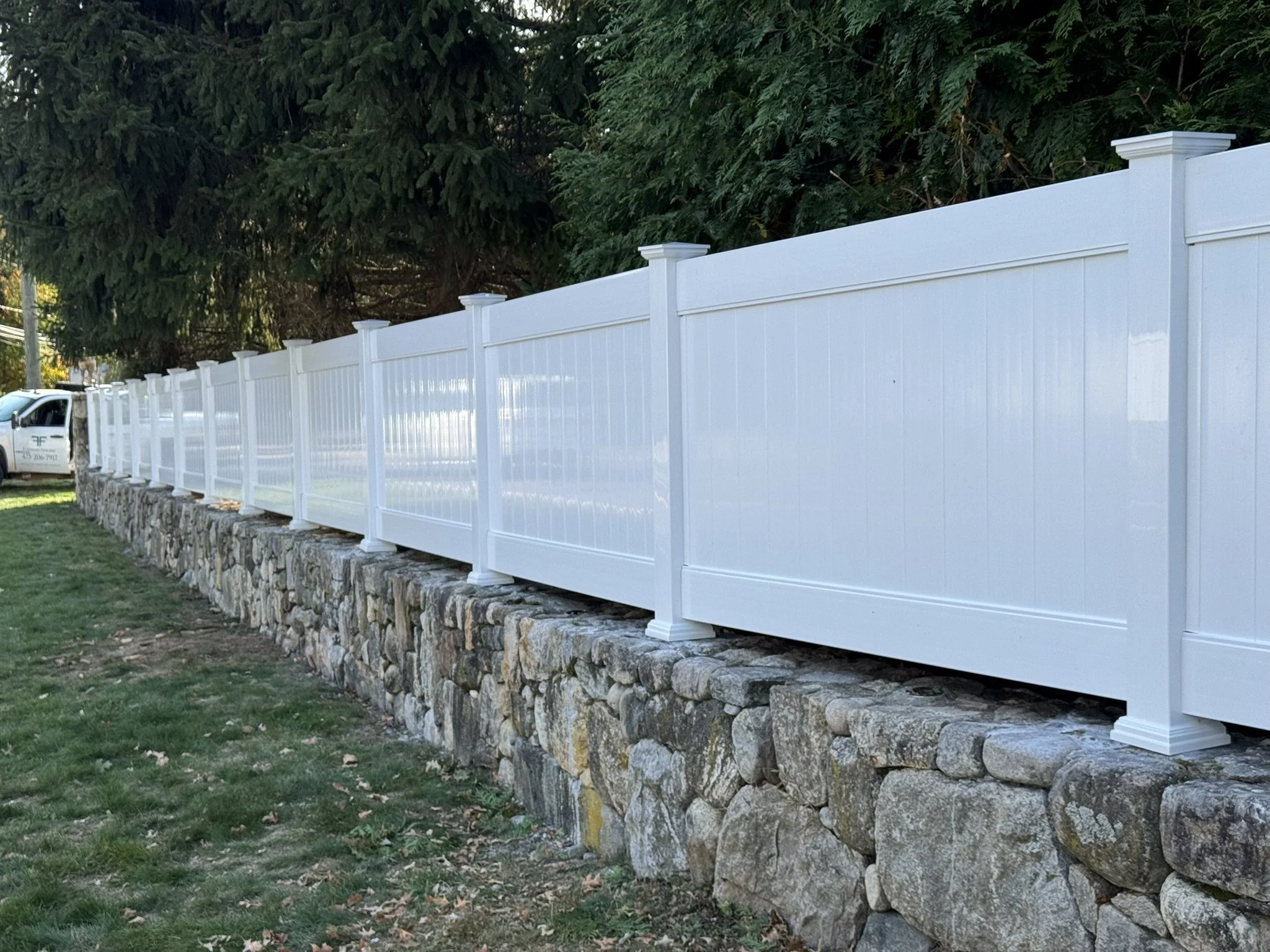 White vinyl fence installed atop a stone wall, with a backdrop of greenery and trees.