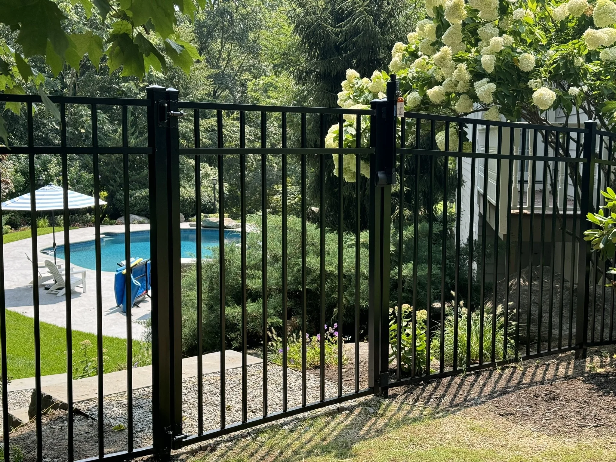 Black metal fence with a gate overlooking a backyard swimming pool area with white chairs, umbrella, and lush greenery.