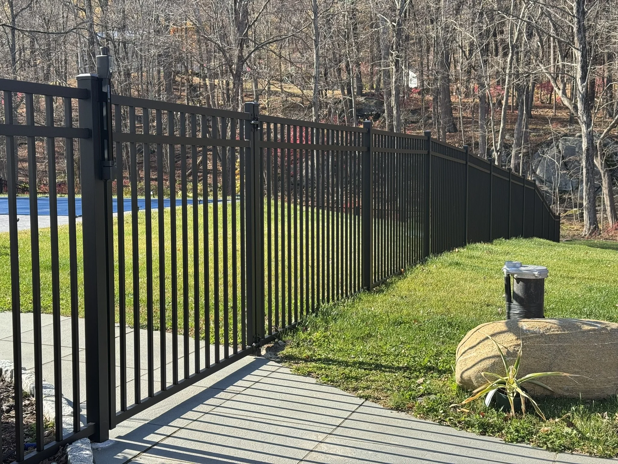 Black metal fence along a sidewalk with a grassy yard, a large rock, a small plant, and a trash can with a lid, with trees and rocks in the background.