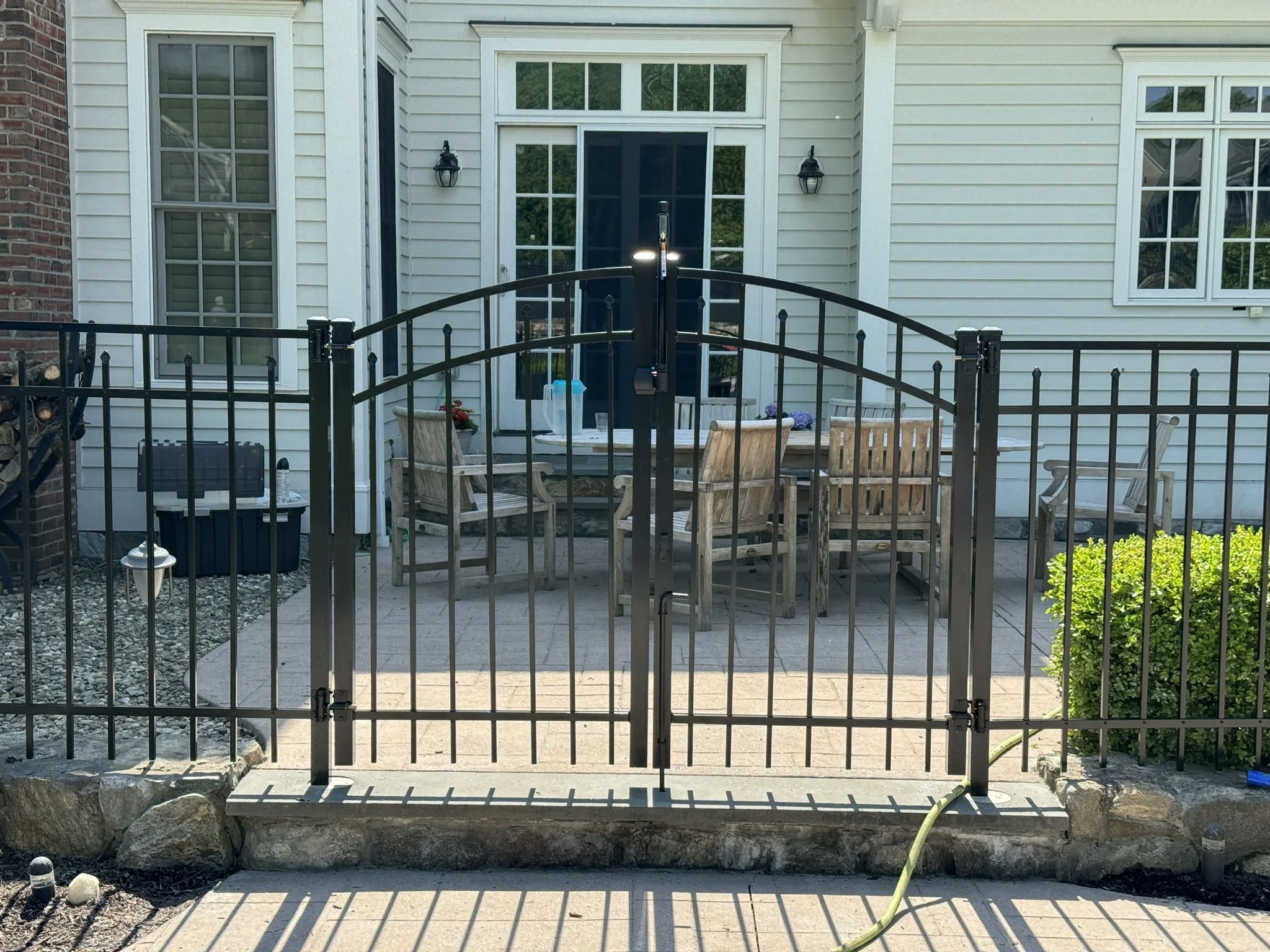 View of a backyard patio with a black metal fence gate, outdoor chairs, a table, and potted plants in front of a white house with beige siding and multiple windows.