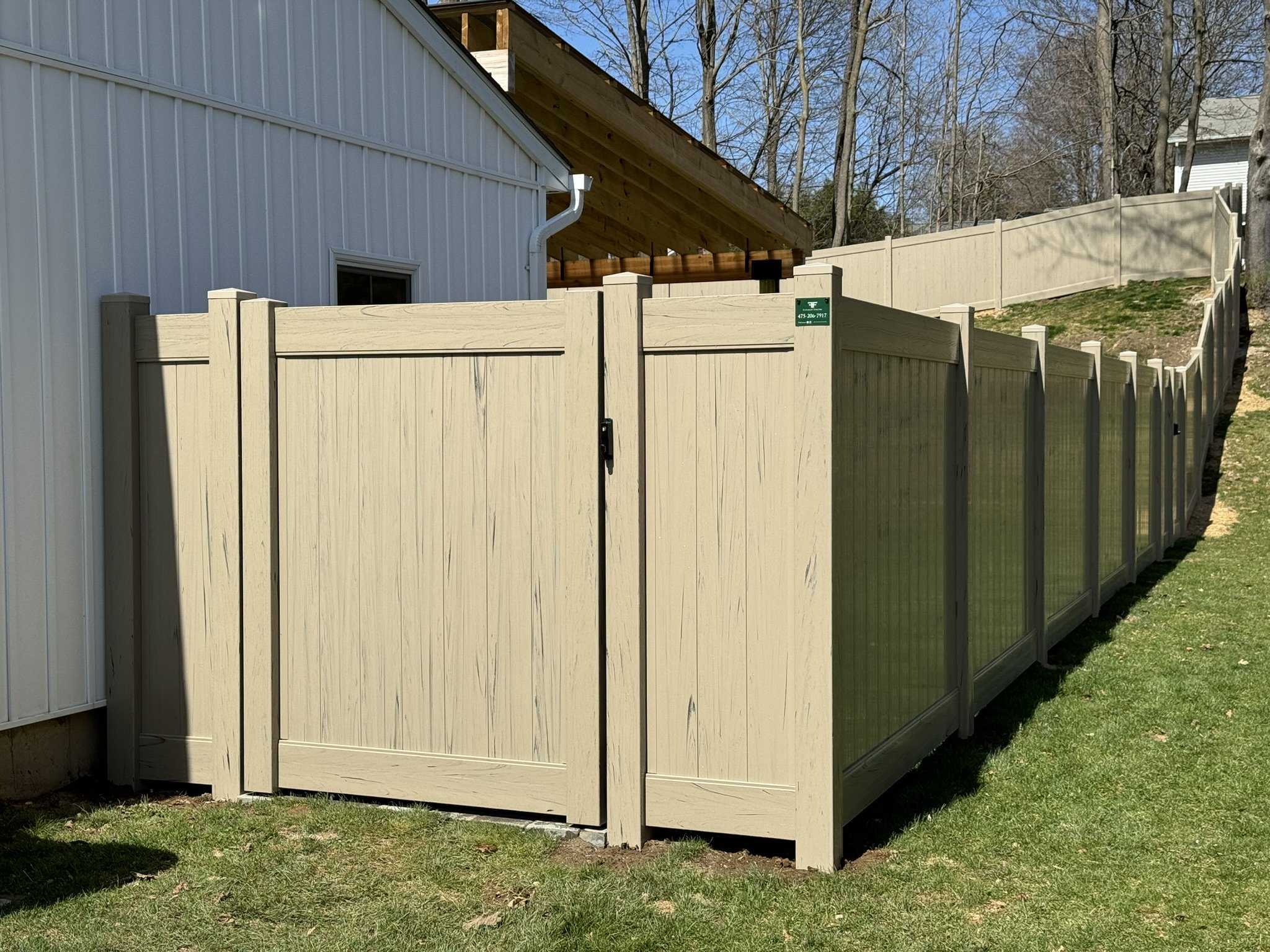 A beige privacy fence around a backyard, with some sections made of solid panels and others with a lattice design. A white building with vertical siding is visible in the background, along with a section of wooden roof under construction.