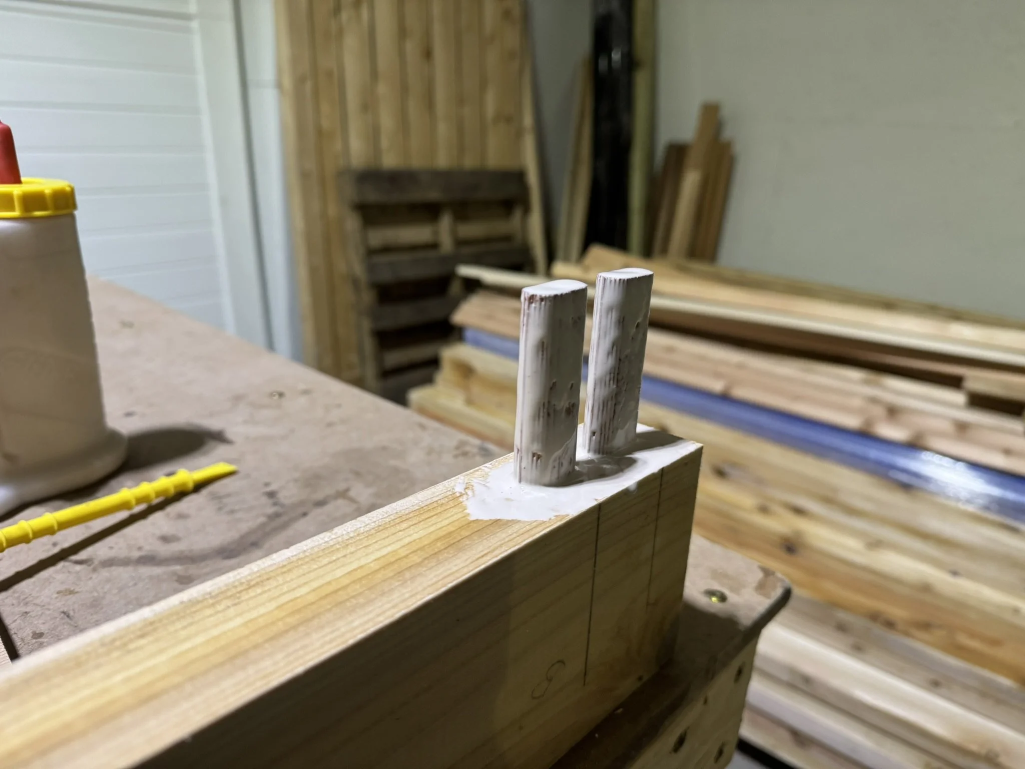 Close-up of a woodworking project with two protruding dowels inserted into a wooden block, with white glue around the joint, with a background of stacked lumber and a storage area.