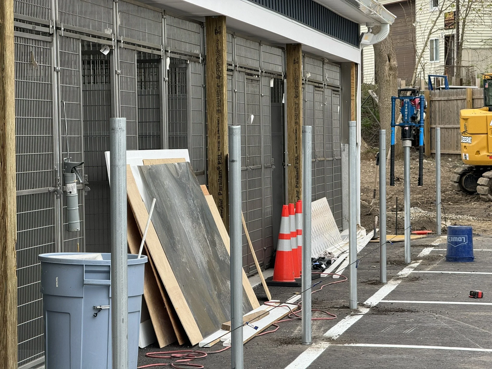 Construction site with metal fence, orange traffic cones, construction tools, and equipment, and a partially built structure, with nearby trees and parked vehicles.