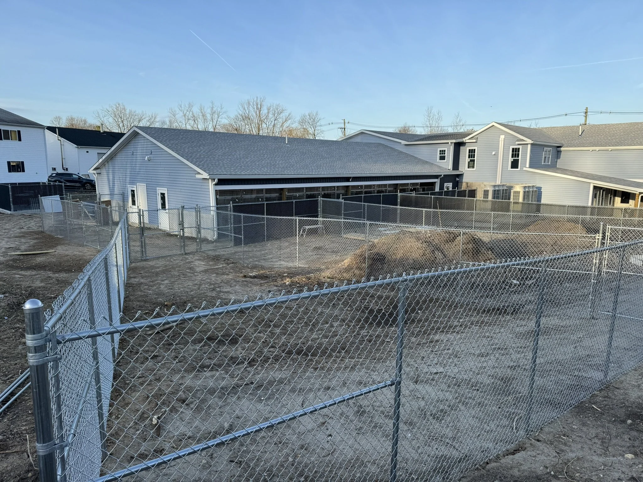 Construction site with dirt piles, surrounded by metal fencing, in front of residential houses with clear blue sky.