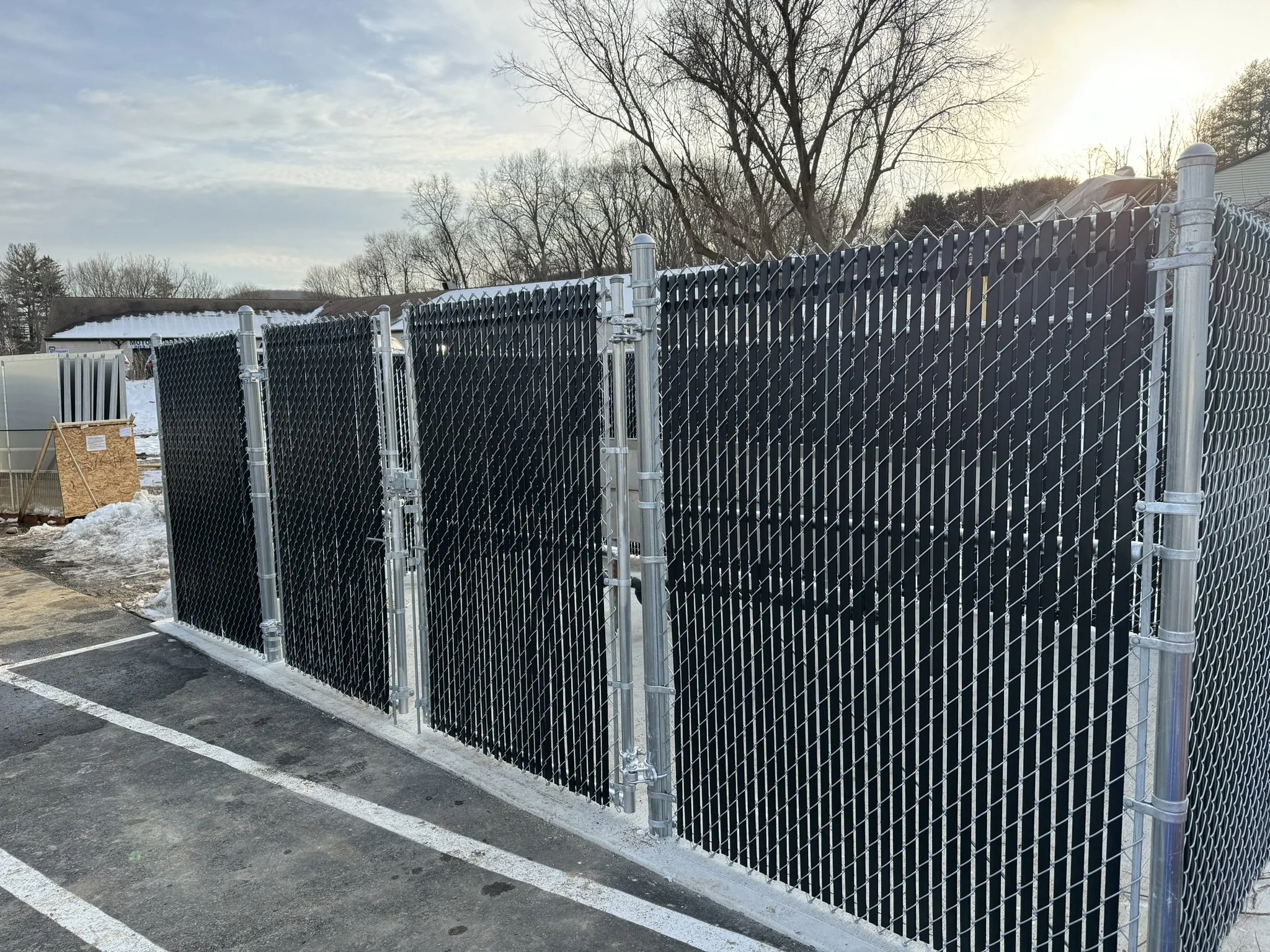 A black metal chain-link fence with metal posts, situated in a parking lot with snow on the ground. In the background, leafless trees and a building are visible under a mostly cloudy sky at sunset.
