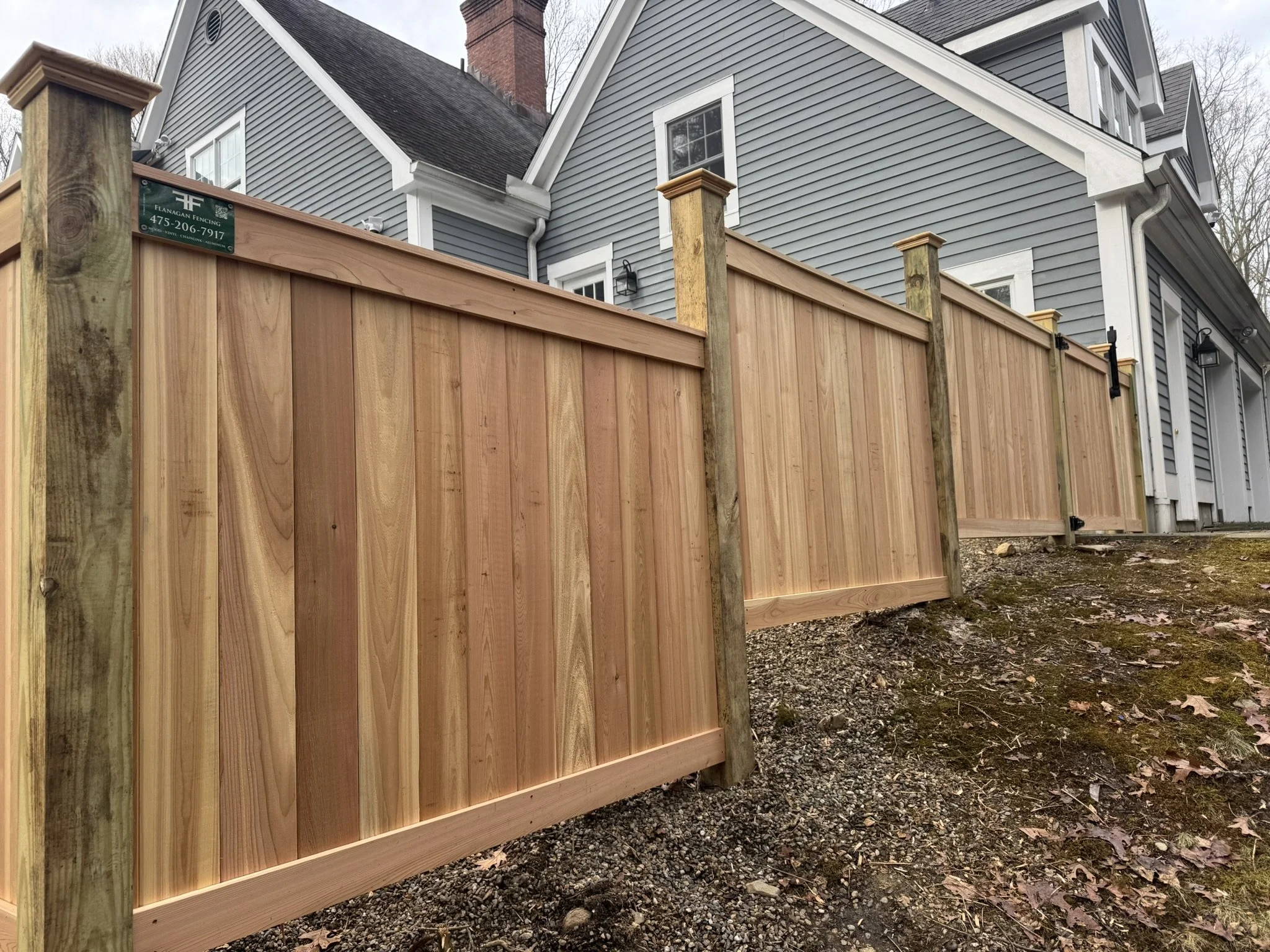 Newly installed wooden privacy fence with vertical planks and support posts in front of gray house with white trim, on sloped ground with fallen leaves.