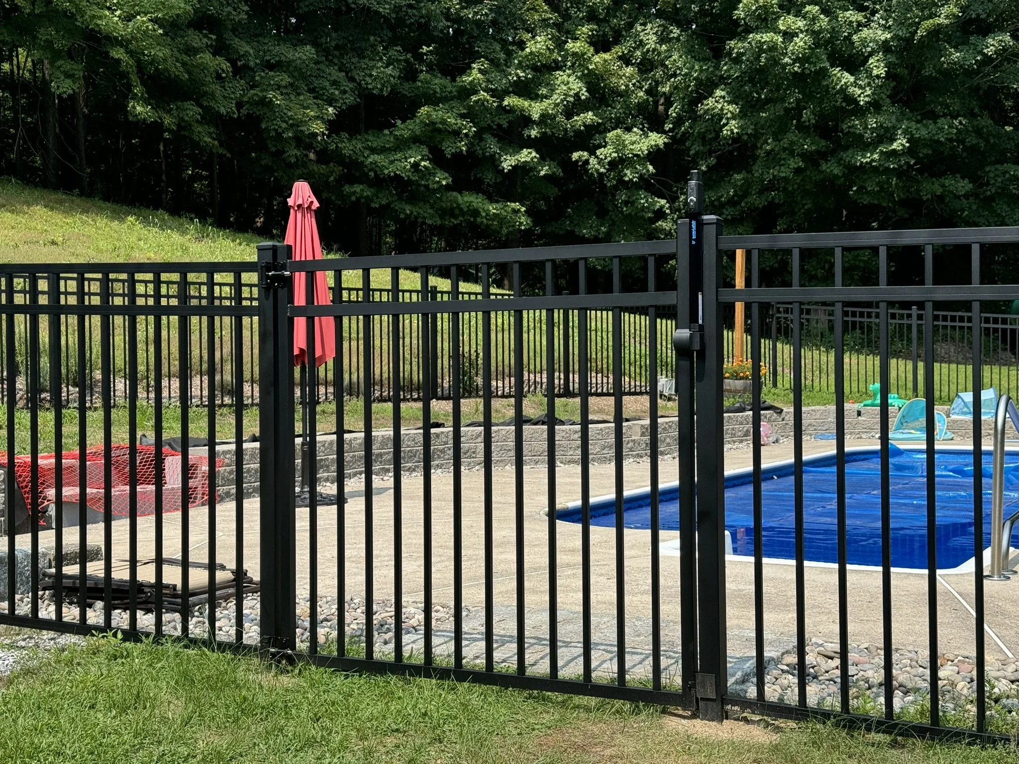 view of a backyard swimming pool with a black metal fence around it, a red patio umbrella, and various pool toys and lounge chairs.