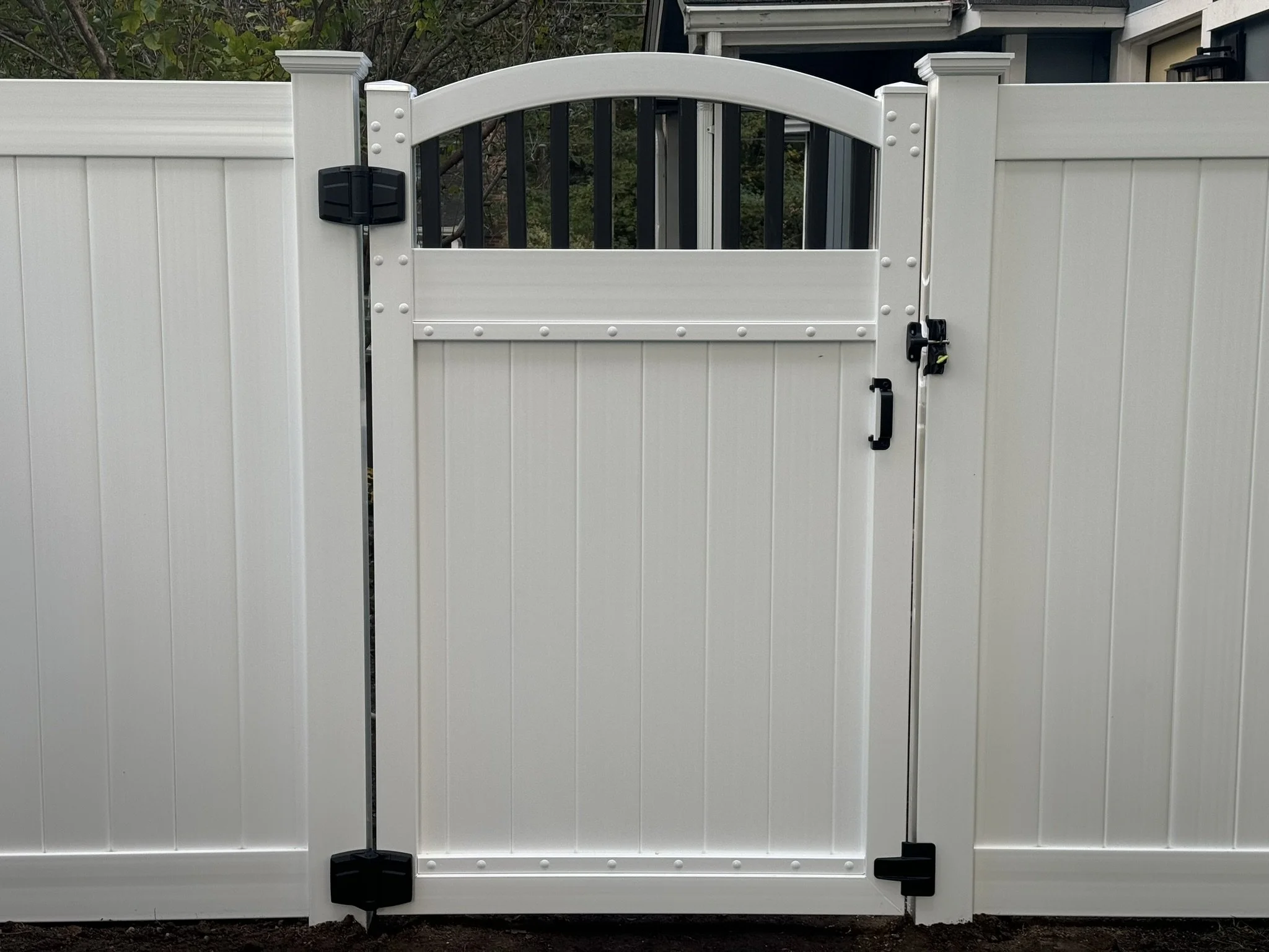 White vinyl fence with a gate secured by black hinges and a latch, featuring an arch on top and ornamental hardware accents.