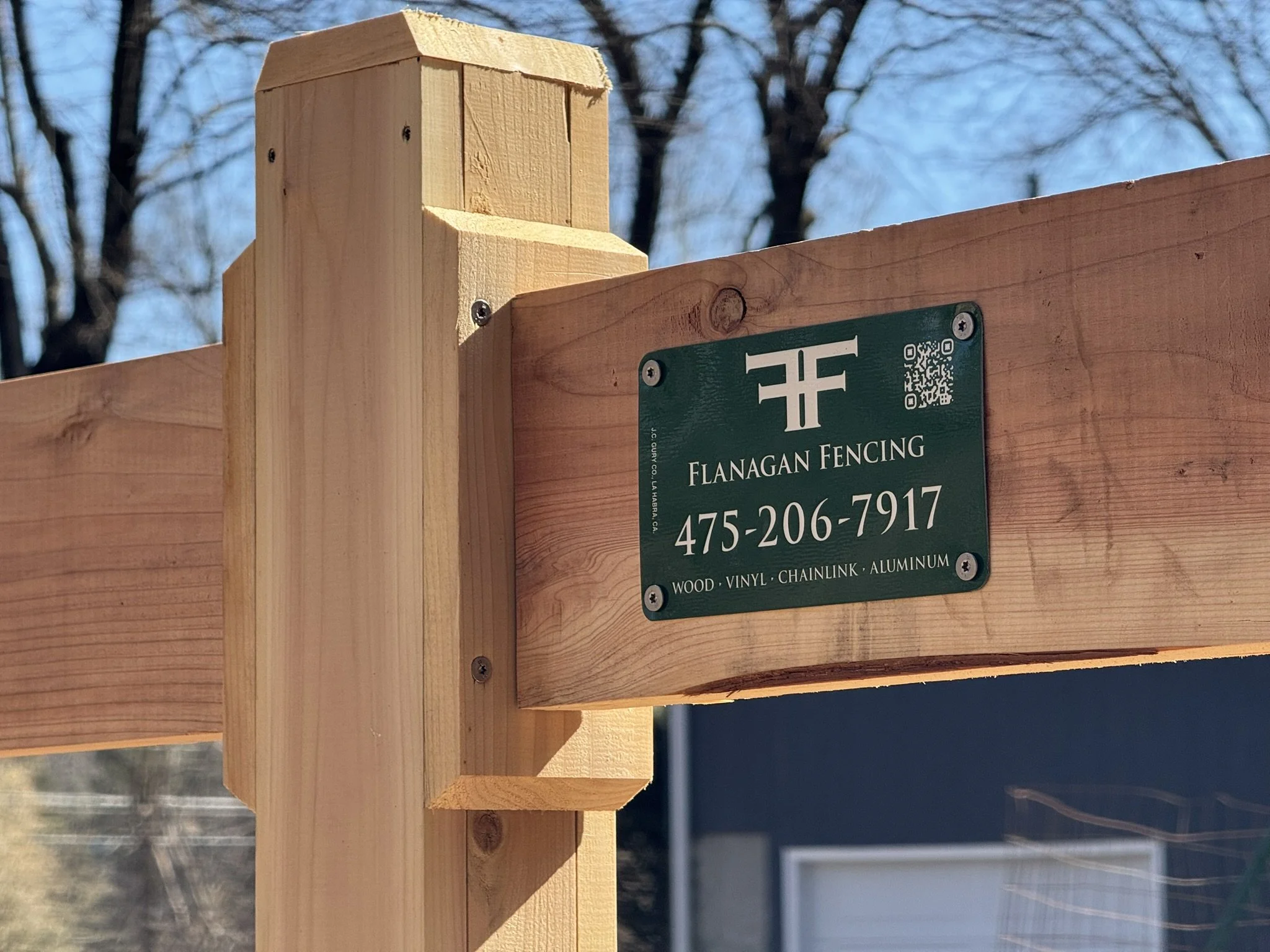 Close-up of a wooden fence post with a green business sign reading 'Flanagan Fencing' and a phone number. The sign mentions materials like wood, vinyl, chainlink, and aluminum.