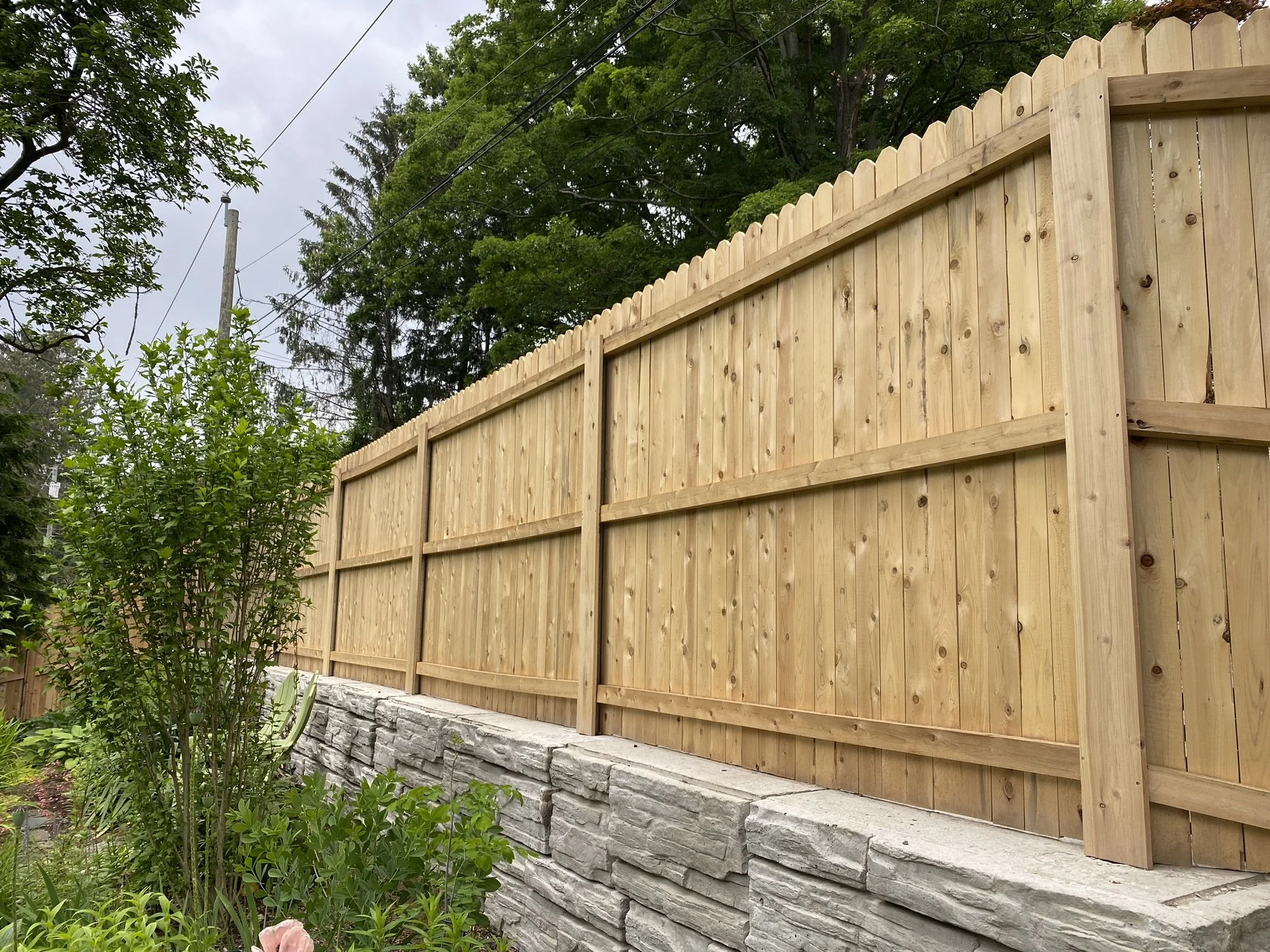 New wooden privacy fence installed on a stone retaining wall in a backyard garden, with trees and power lines in the background.