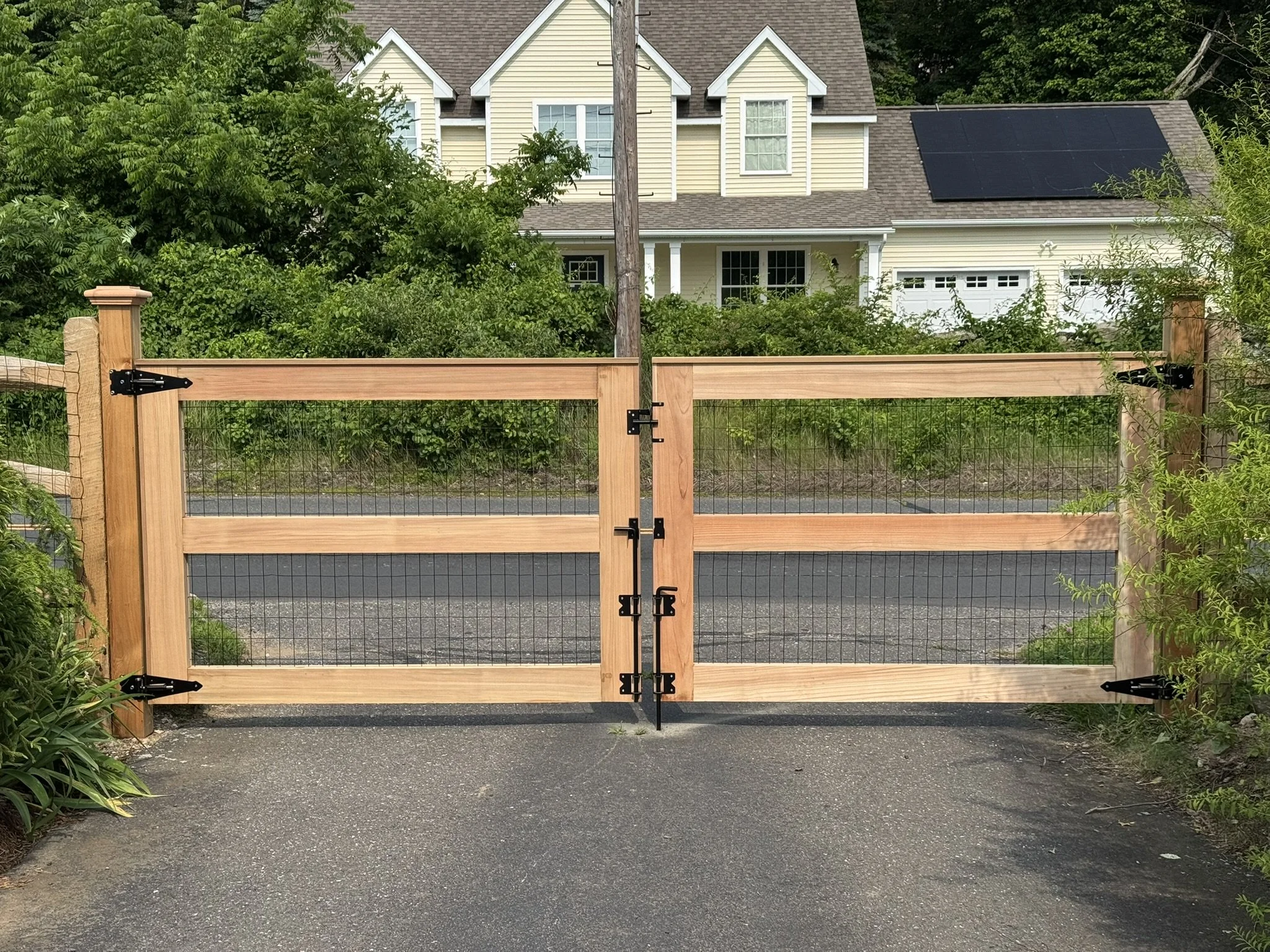 Wooden and wire mesh driveway gate with black hinges, opening to a house with solar panels on the roof, surrounded by greenery.