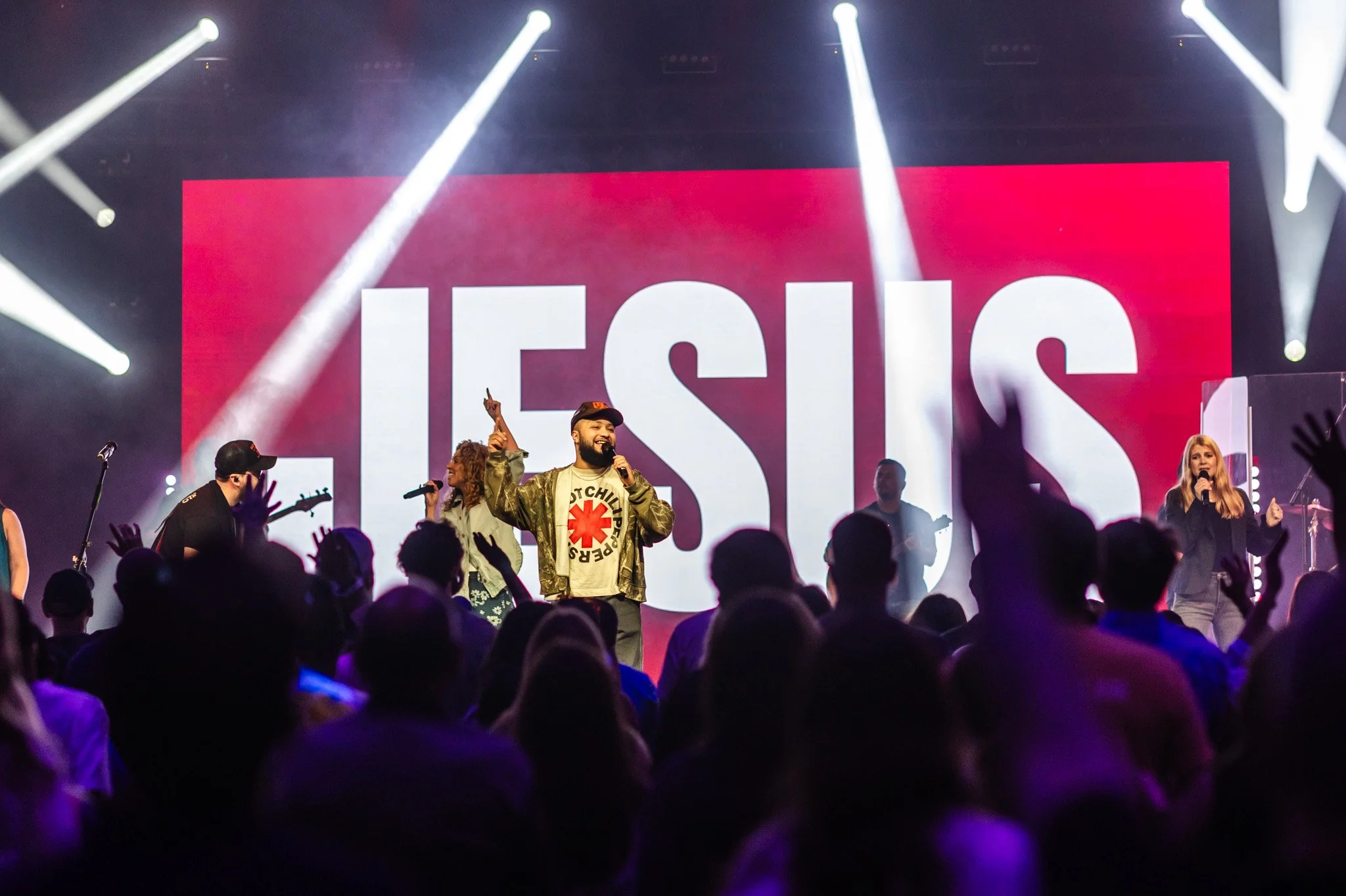 A group of performers on stage during a religious event with a large screen behind them displaying the word 'JESUS' in bold letters. Audience members are in the foreground, some raising their hands.
