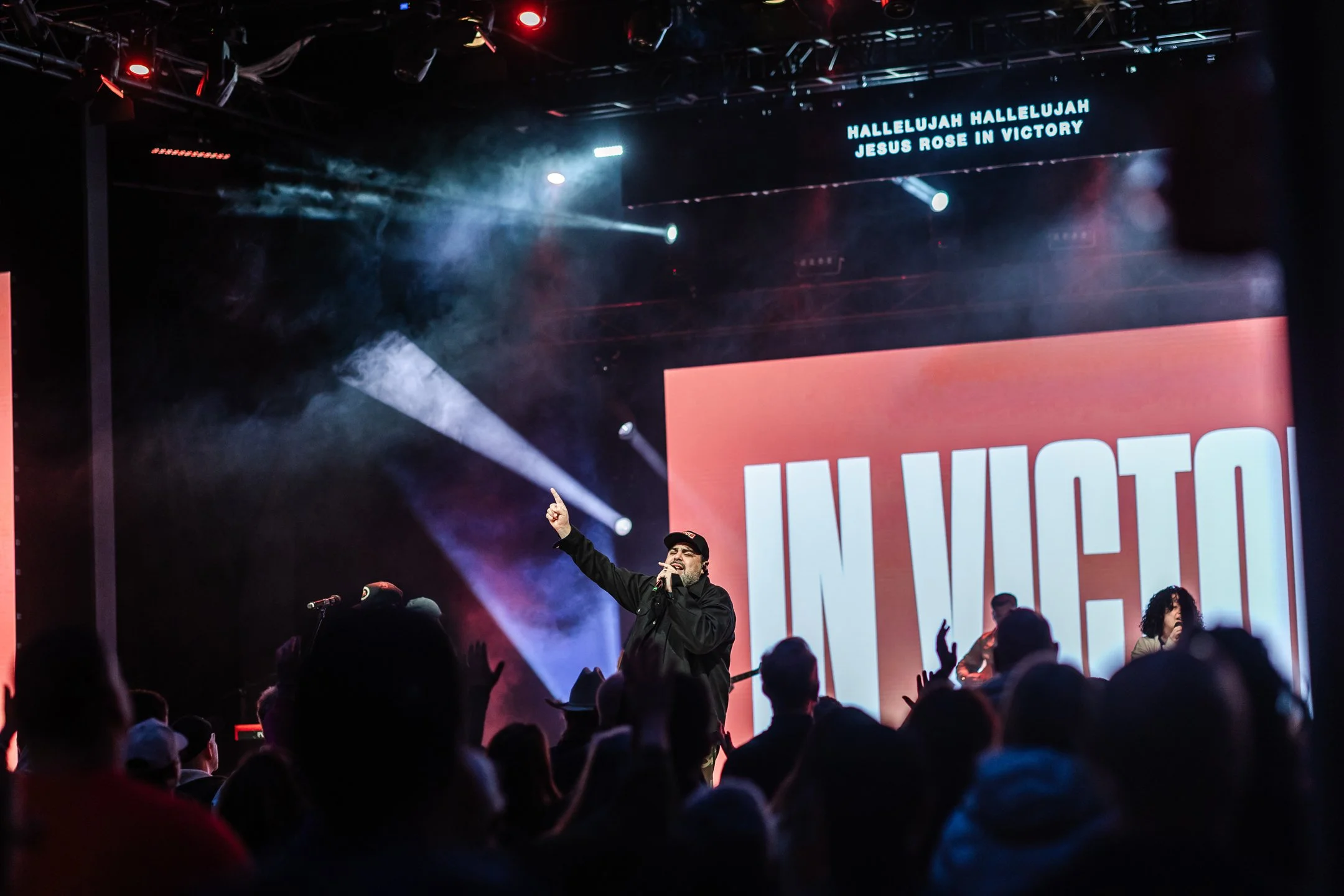 A performer singing on stage with a microphone in hand, raising one finger, in front of an audience. The stage has colorful lights and a large screen displaying the words 'IN VICTO' and text above that says 'HALLELUJAH HALLELUJAH JESUS ROSE IN VICTORY'.