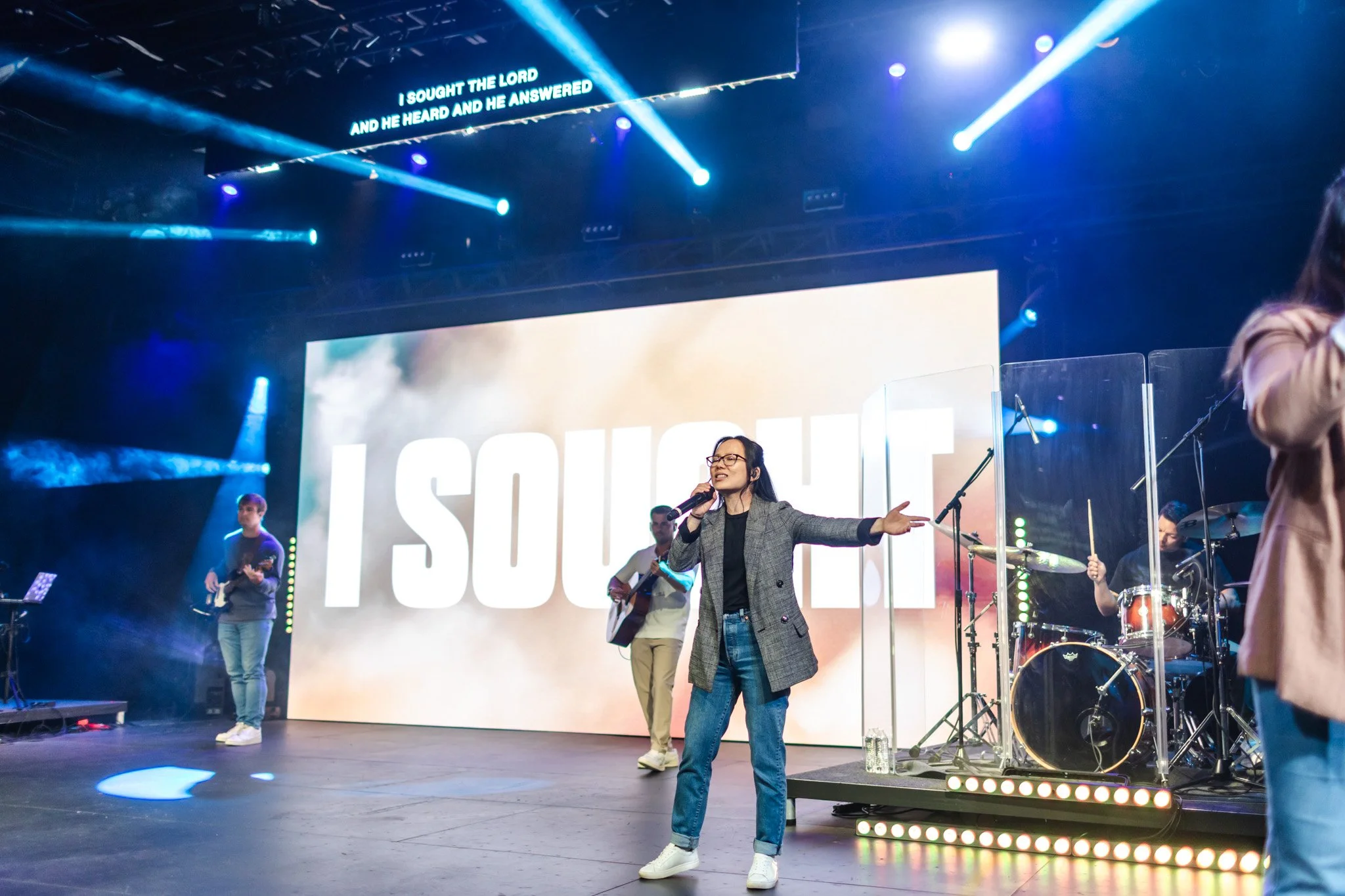 Woman singing on stage with a band, large screen behind her displaying the word 'I SOUGHT,' and stage lights shining down.