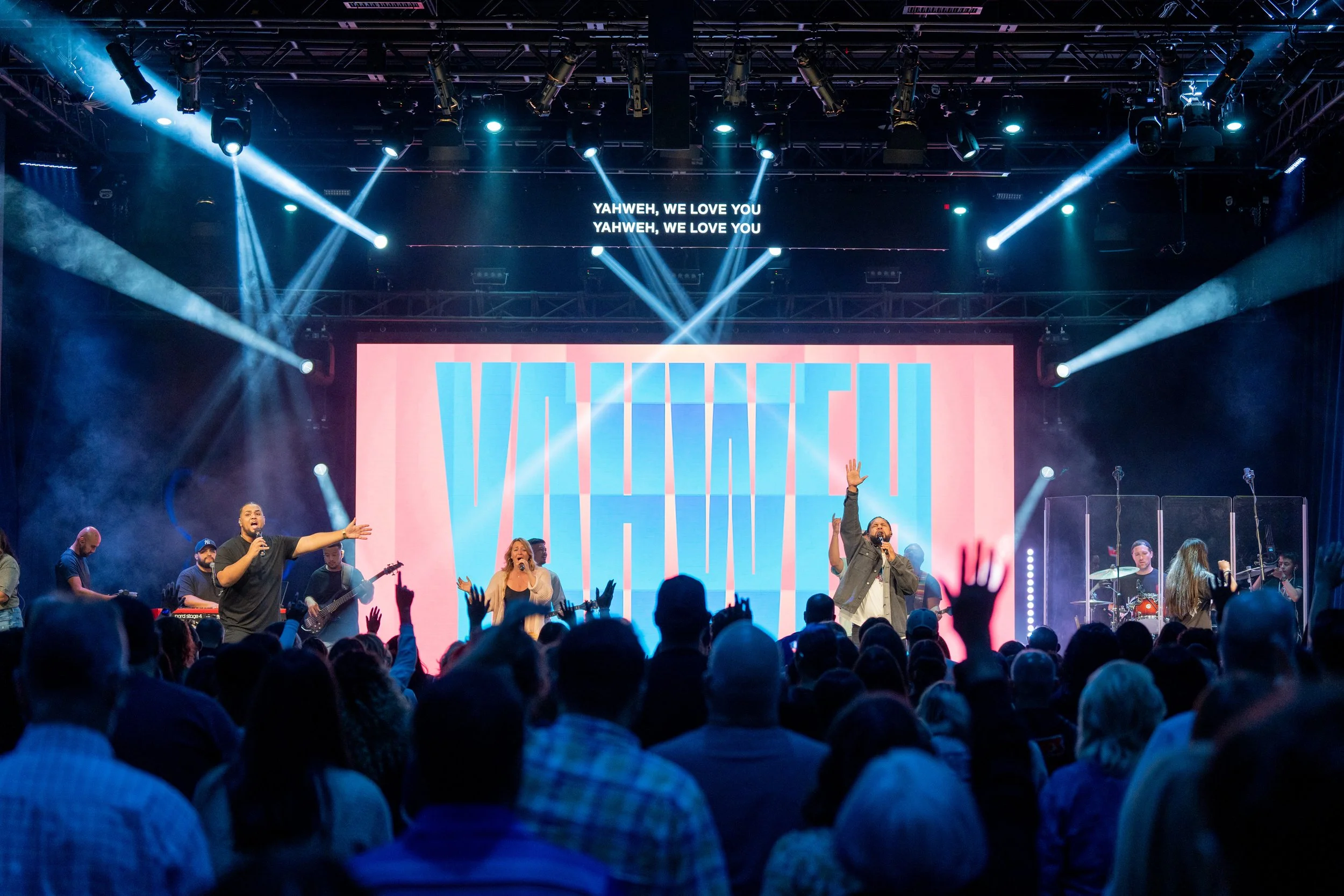 A large crowd attending a live worship concert with a stage, worship band, and singers. The stage features a big screen displaying the words 'VICTORY' with vibrant lighting and spotlights. Lyrics, 'YAHWEH, WE LOVE YOU,' are shown on a smaller screen above.