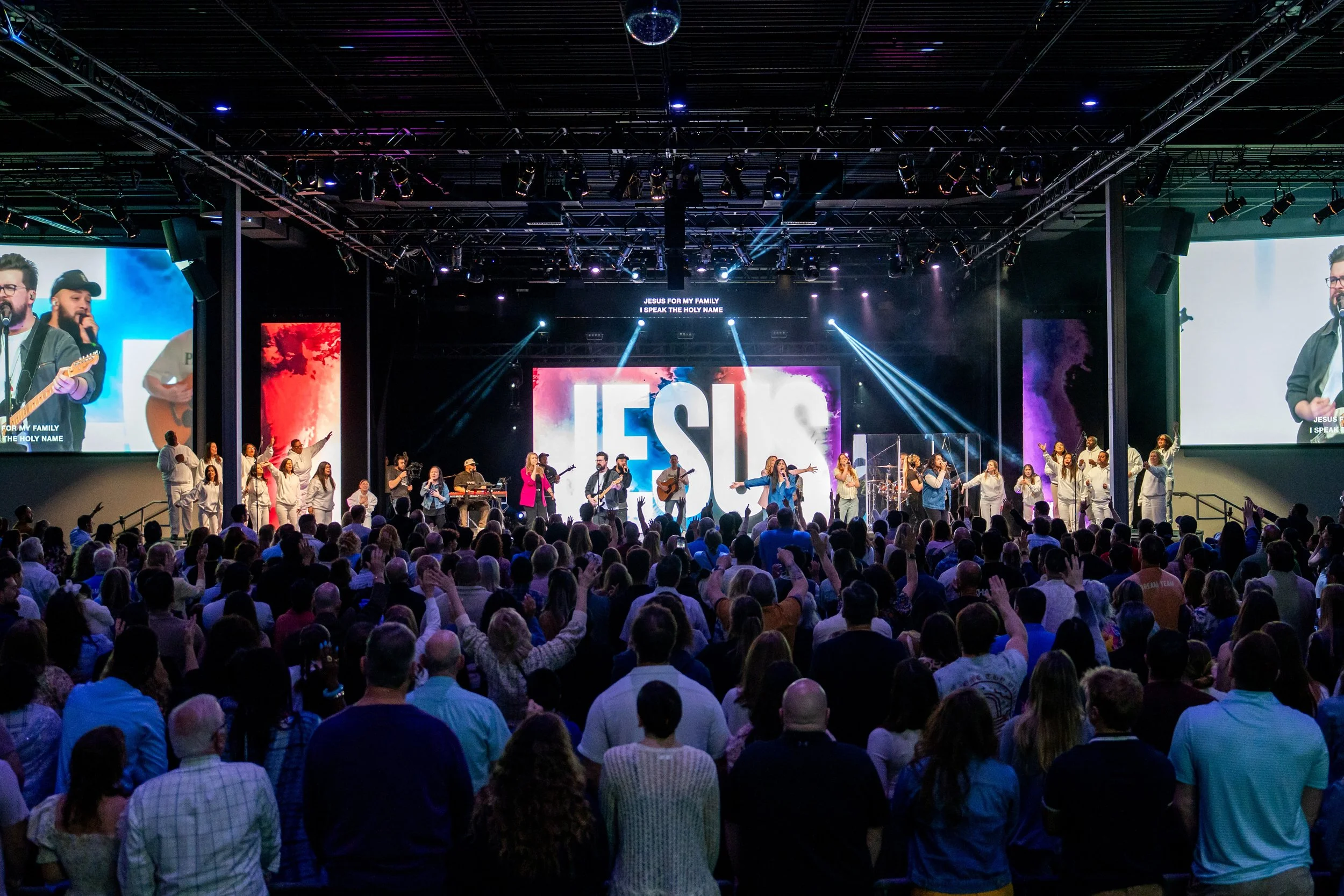 A large congregation attending a church service with musicians and singers performing on a stage, large screens display the word 'JESUS' in the background, with people raising their hands in worship.