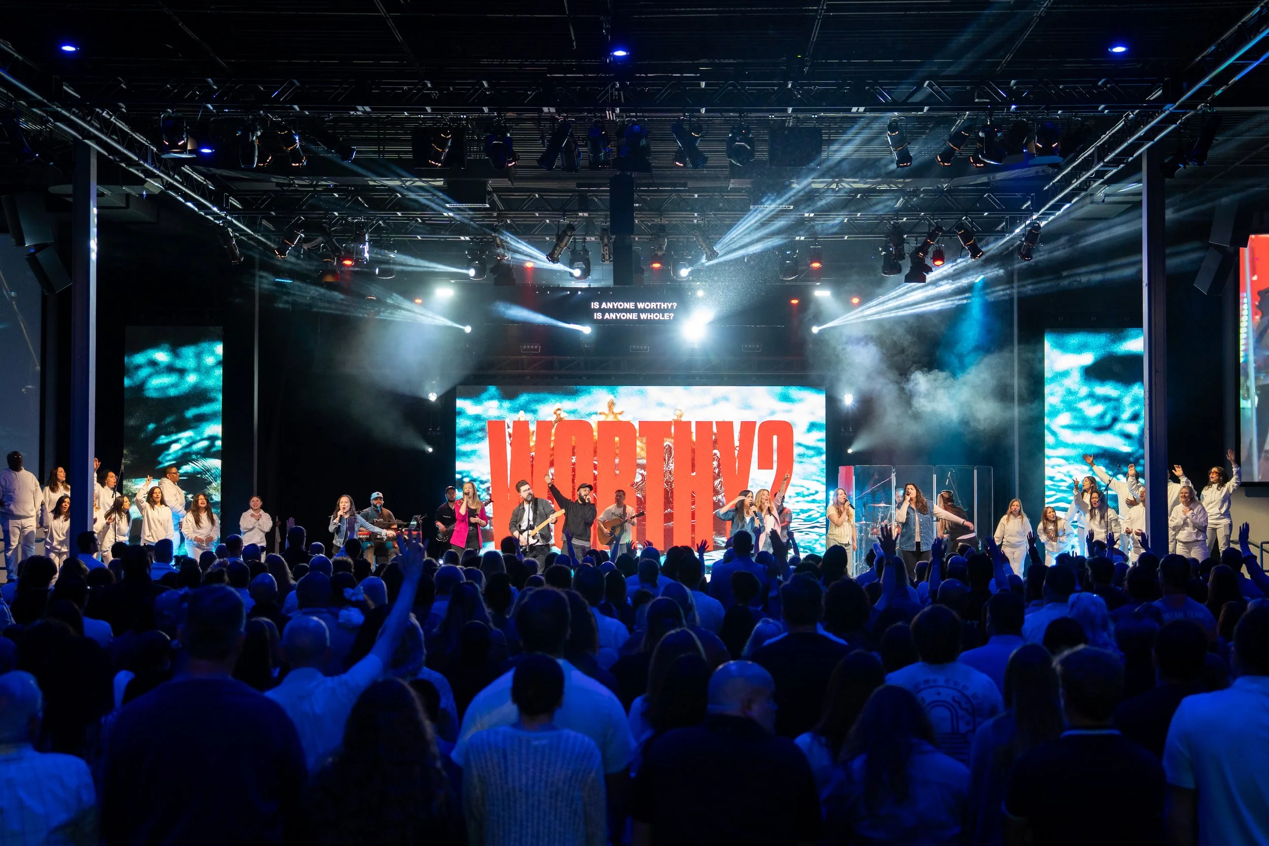 A concert or worship event on a large stage with musical performers, singers, and dancers, with a large audience watching and raising their hands. The stage has bright lights, smoke effects, and a digital screen with the word 'WORTHY' displayed in red and white.