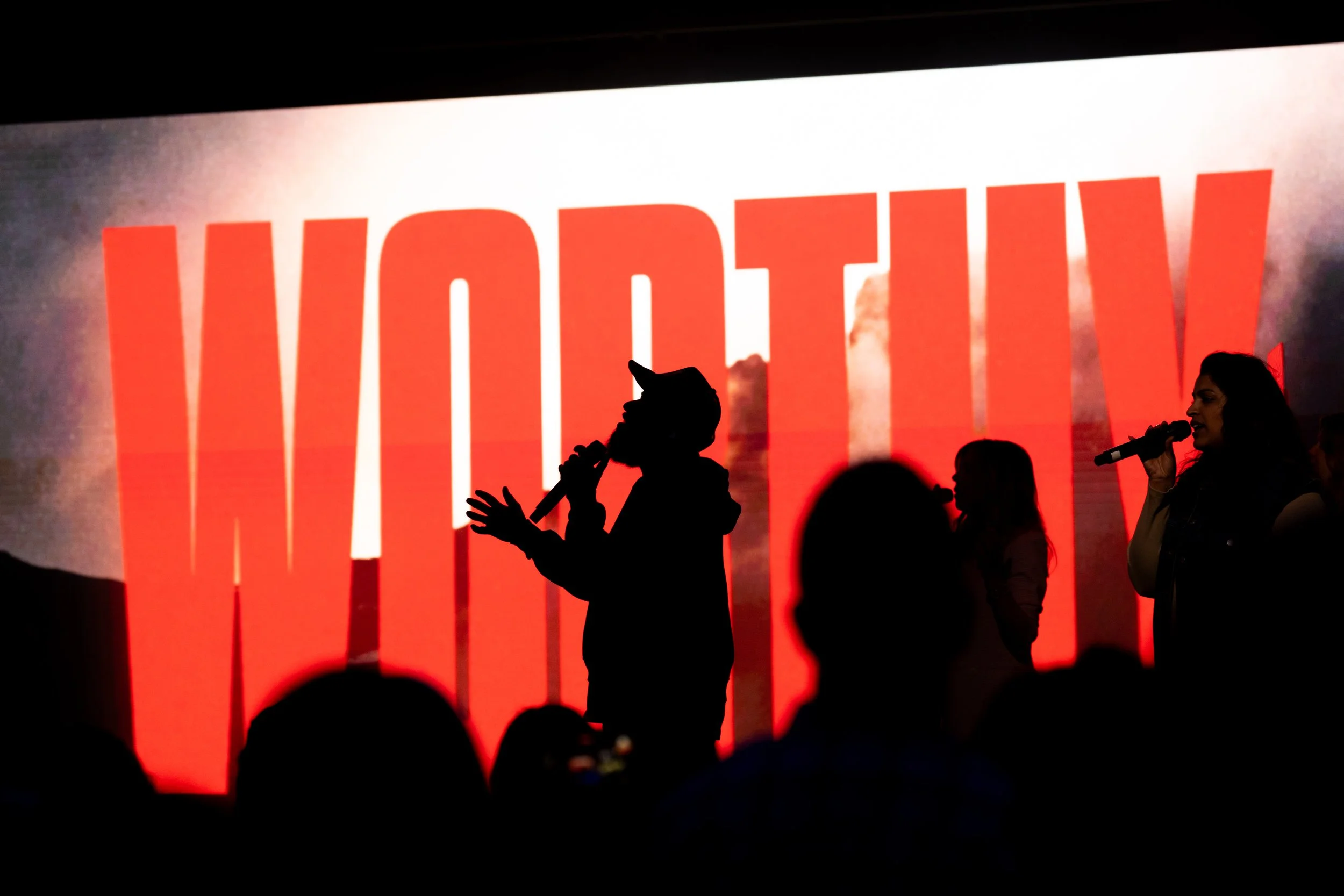 Silhouettes of people speaking or singing into microphones on stage with a large, illuminated 'WONDER WOMAN' sign in the background.
