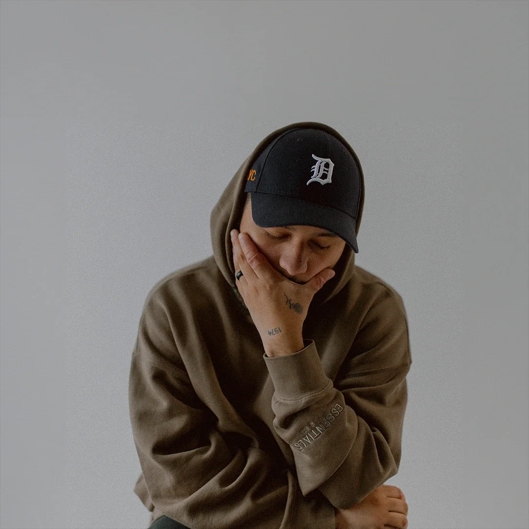 A person wearing a brown hoodie and a black baseball cap with a white 'D' logo, resting their chin on their hand with a thoughtful expression.