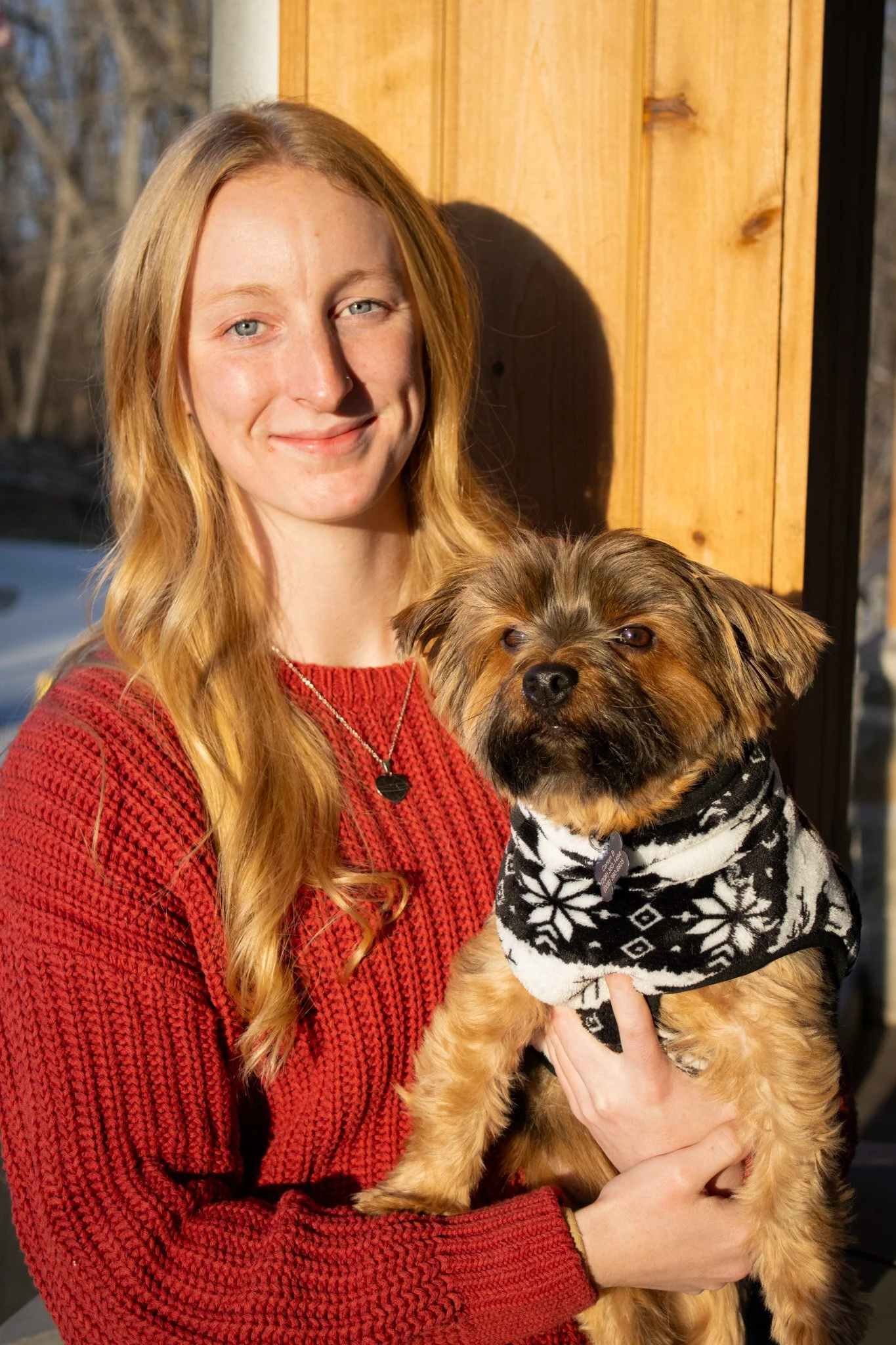 A young woman with long blonde hair in a red sweater holding a small brown dog wearing a black and white sweater, outdoors near a wooden structure.
